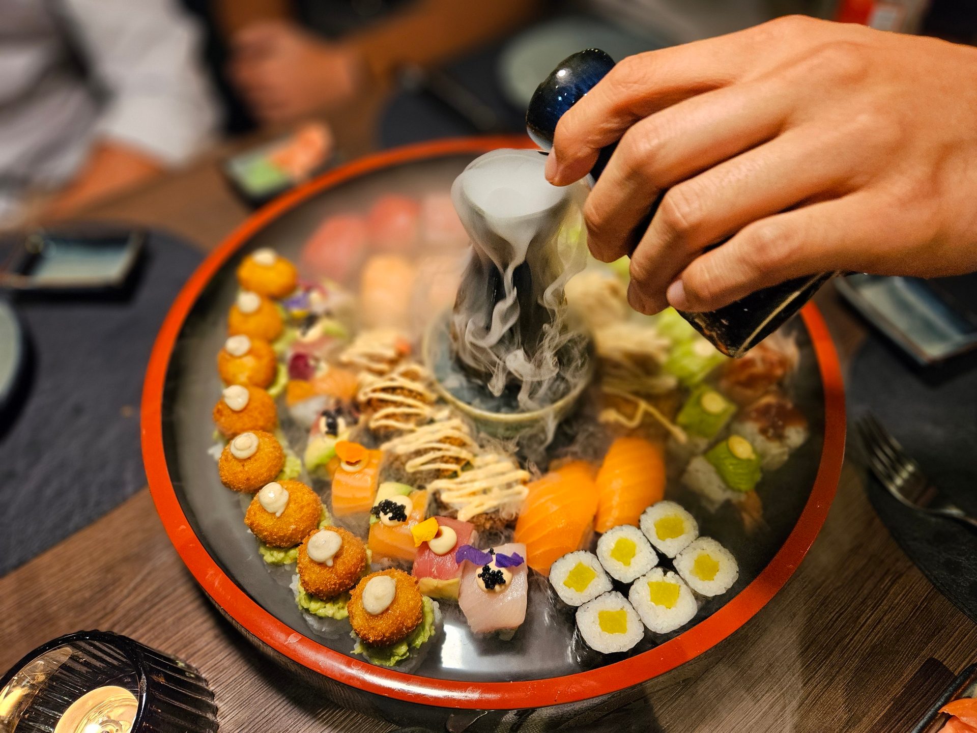 Hand pouring liquid into a smoking sushi platter with various sushi rolls.