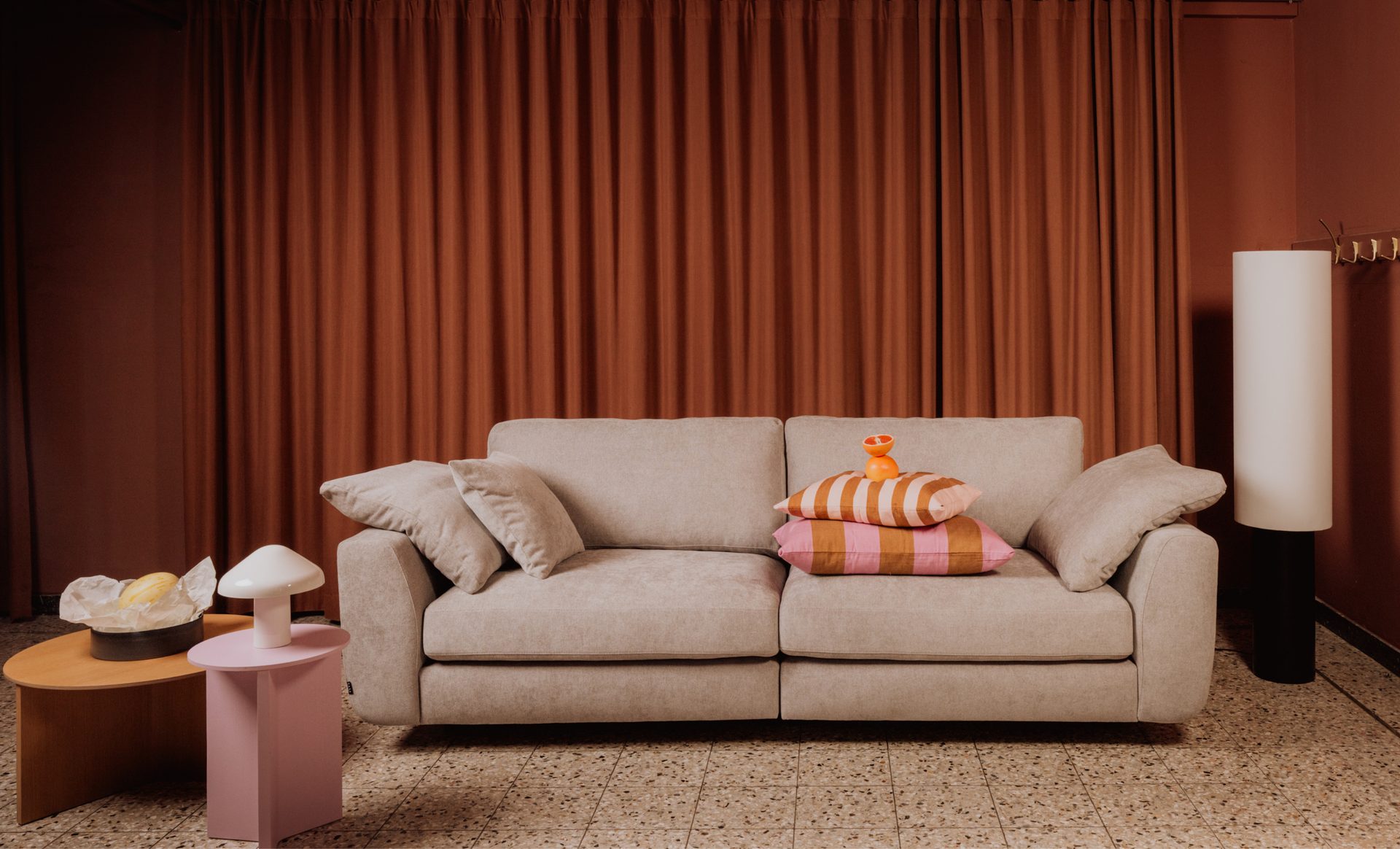 A stylish beige sofa with colorful pillows and modern tables in a room with orange curtains.