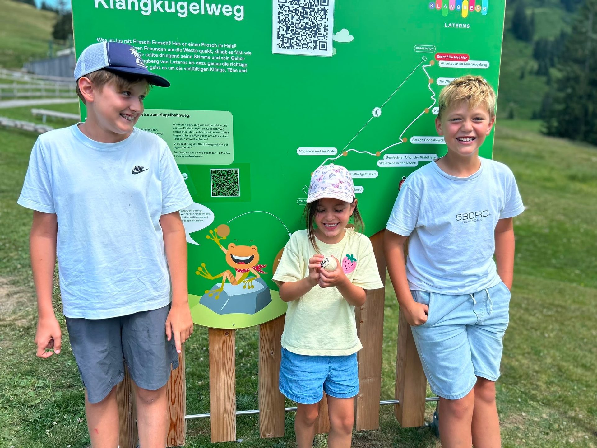 Three smiling children, two boys and one girl, standing in front of a green sign for a 'Klangkugelweg'.