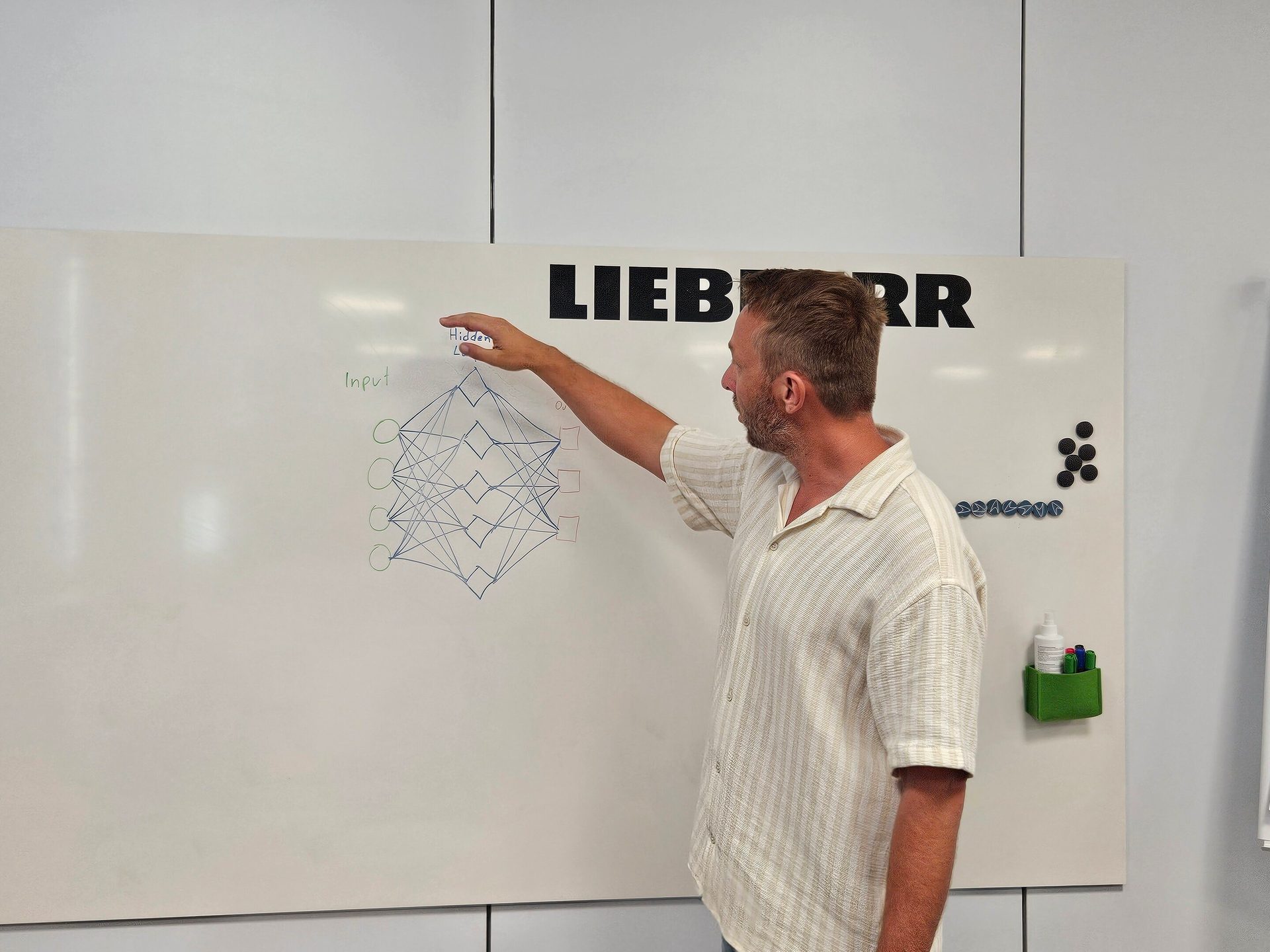 Man pointing at a neural network diagram on a whiteboard with a LIEBHERR logo.