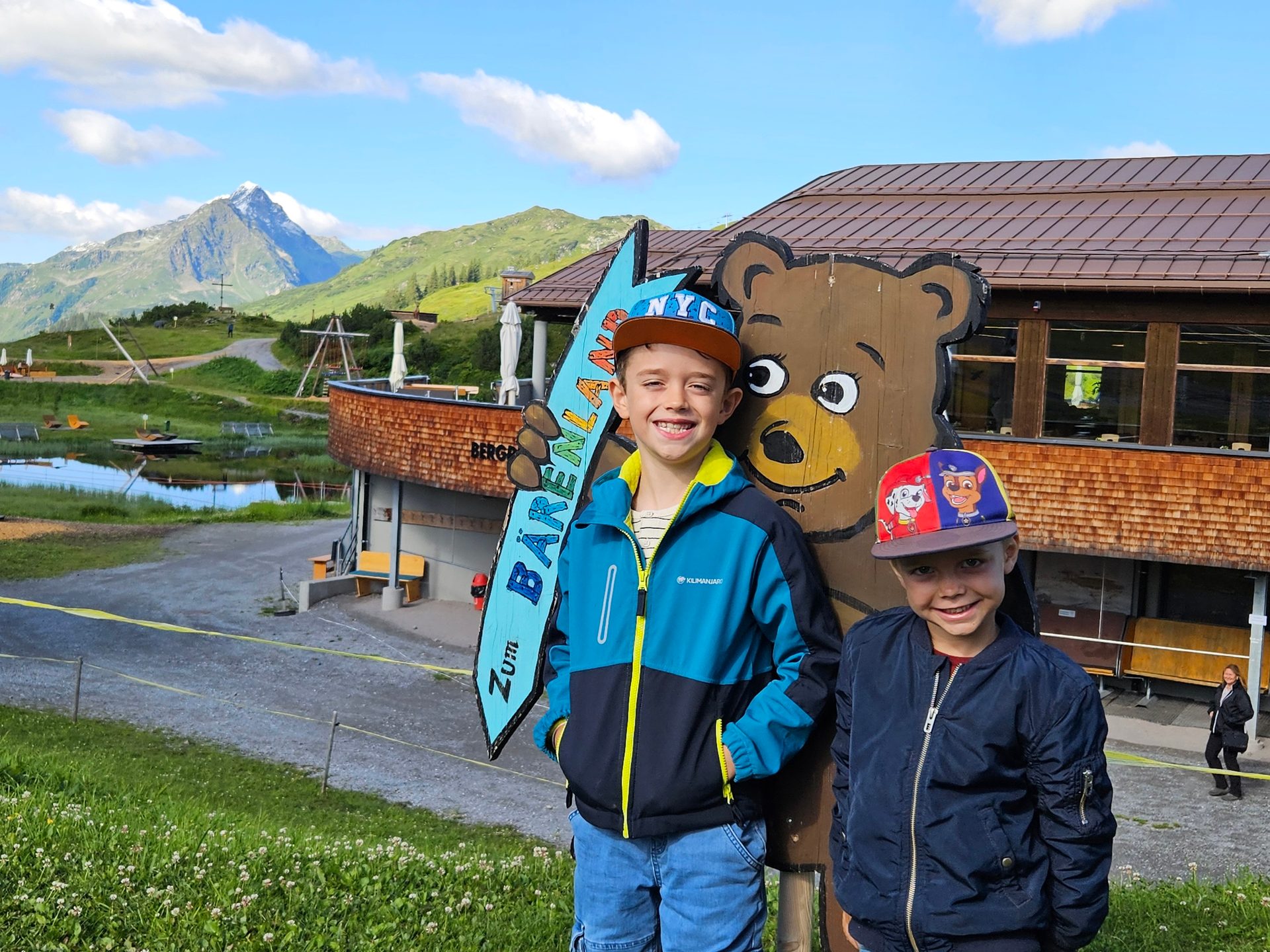 Two smiling boys with a bear cutout, mountains, a building, and a lake in the background.
