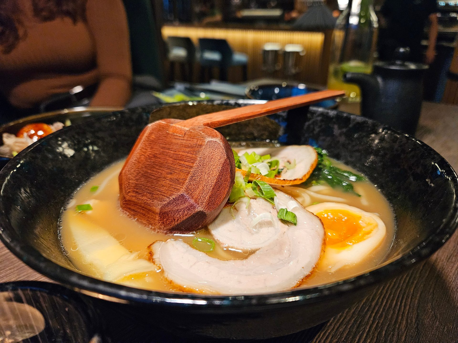 A black bowl of ramen with chashu, soft-boiled egg, scallions, and a wooden ladle in a restaurant.