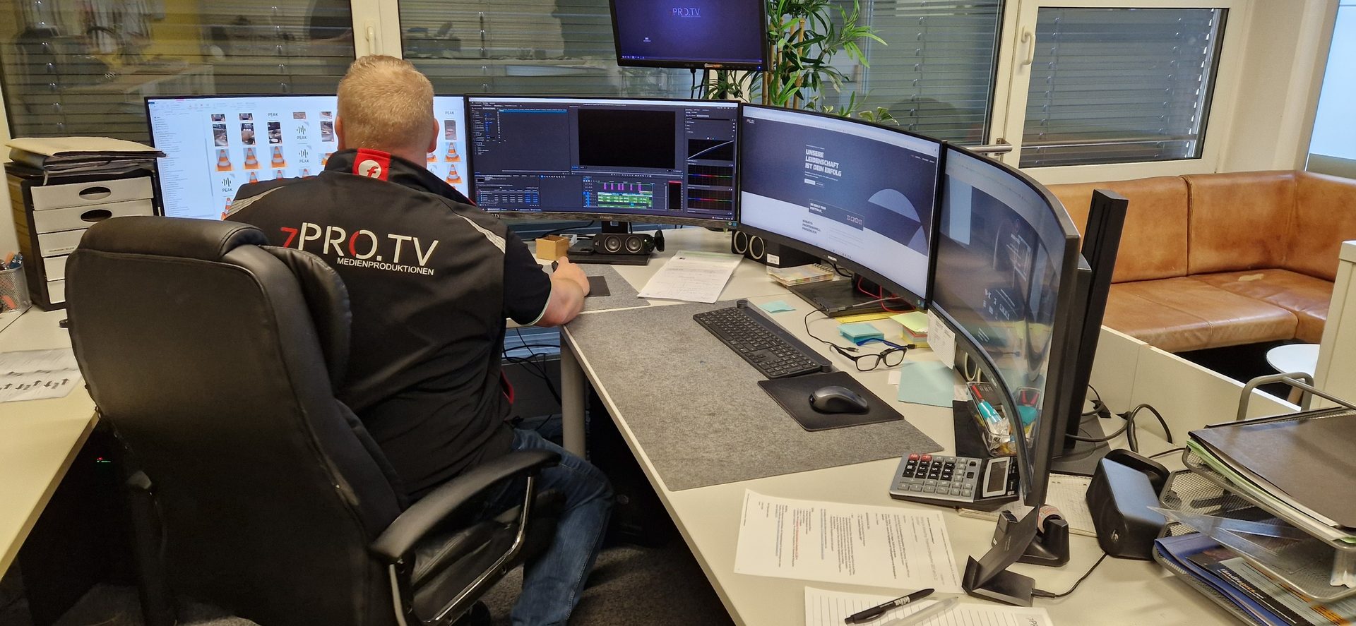 Man in "7PRO.TV" shirt at a multi-monitor computer workstation in an office.