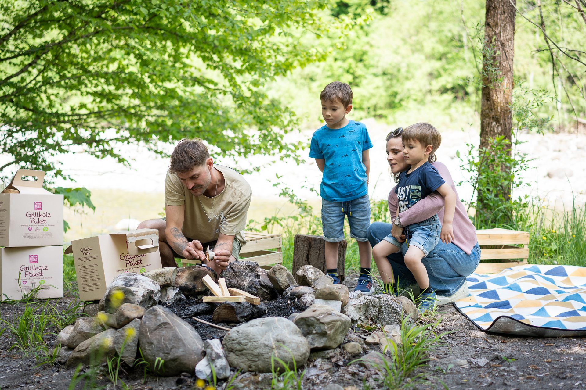 Family lights a campfire outdoors with Grillholz Paket boxes, children watch.