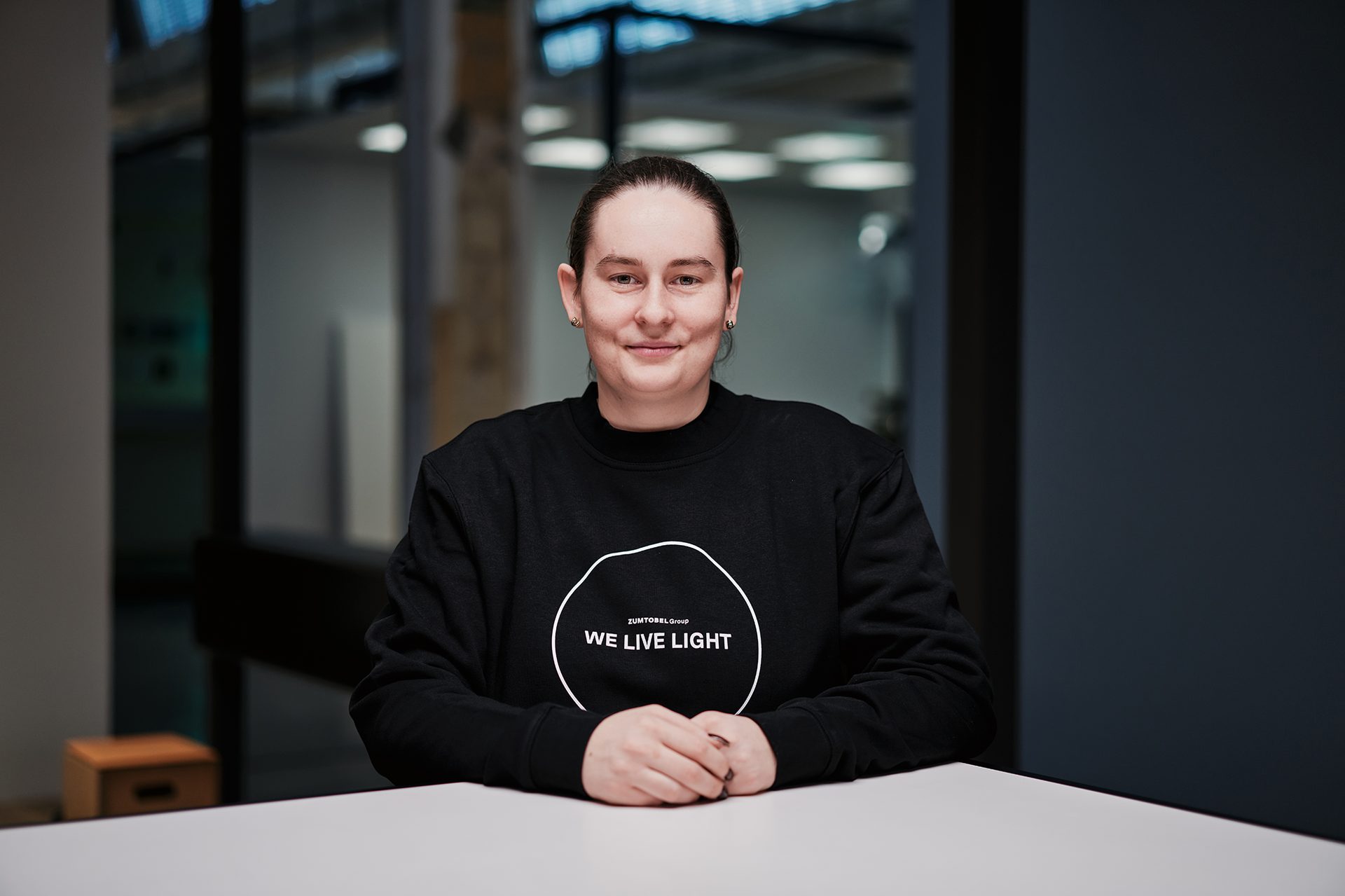 A smiling person in a black "WE LIVE LIGHT" sweatshirt sits at a table.
