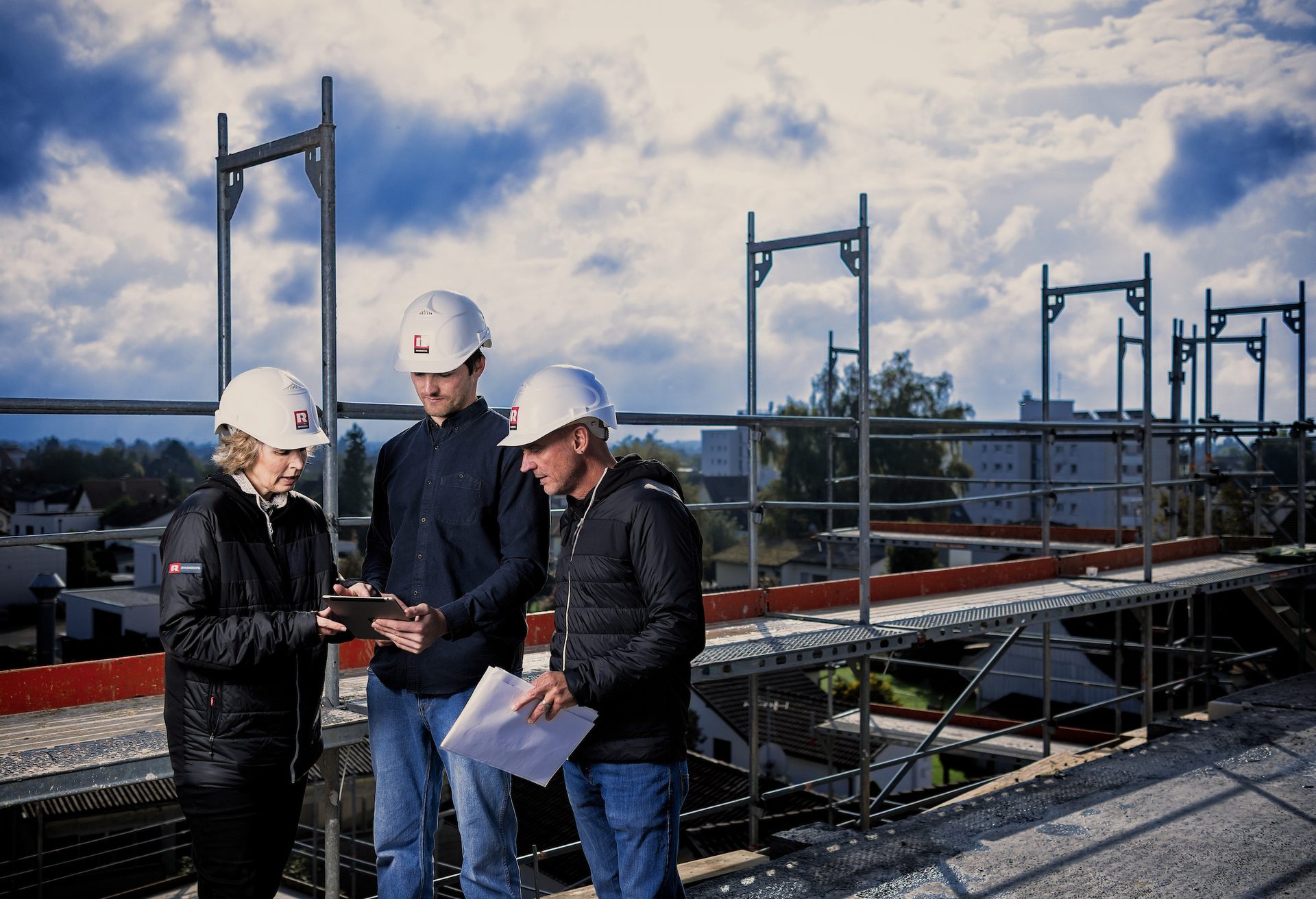 Three construction workers in hard hats on scaffolding reviewing a tablet.