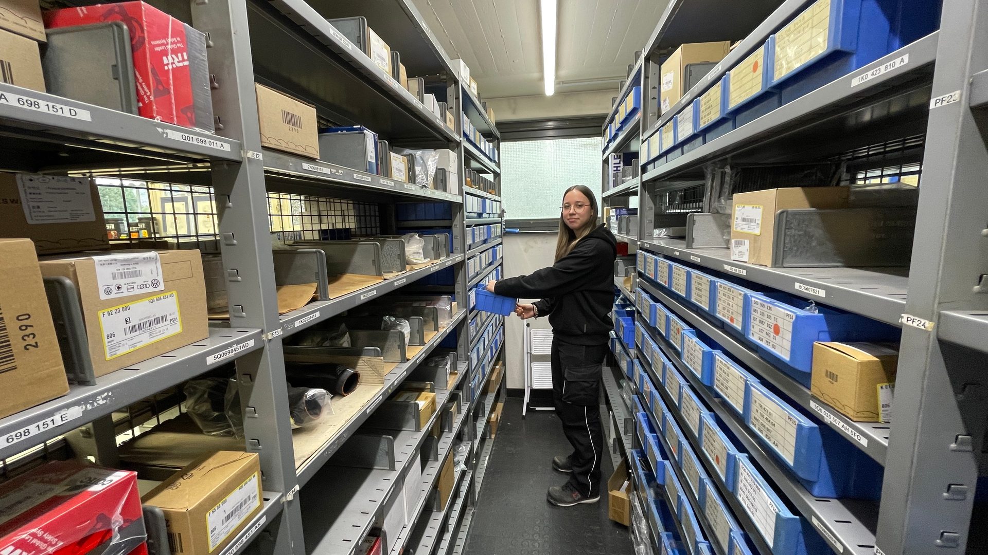 A woman holding a blue bin stands in a narrow warehouse aisle flanked by tall shelves stocked with boxes and parts.