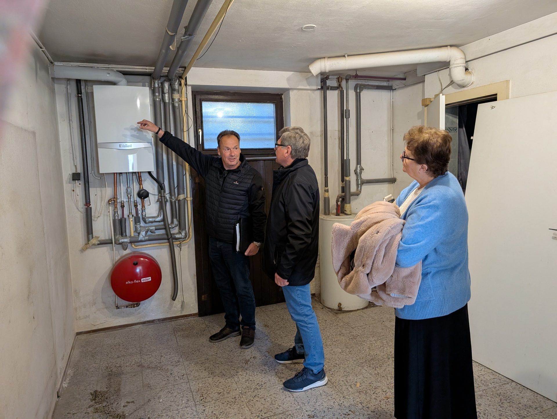Three people in a utility room, one man pointing at a white wall-mounted boiler.