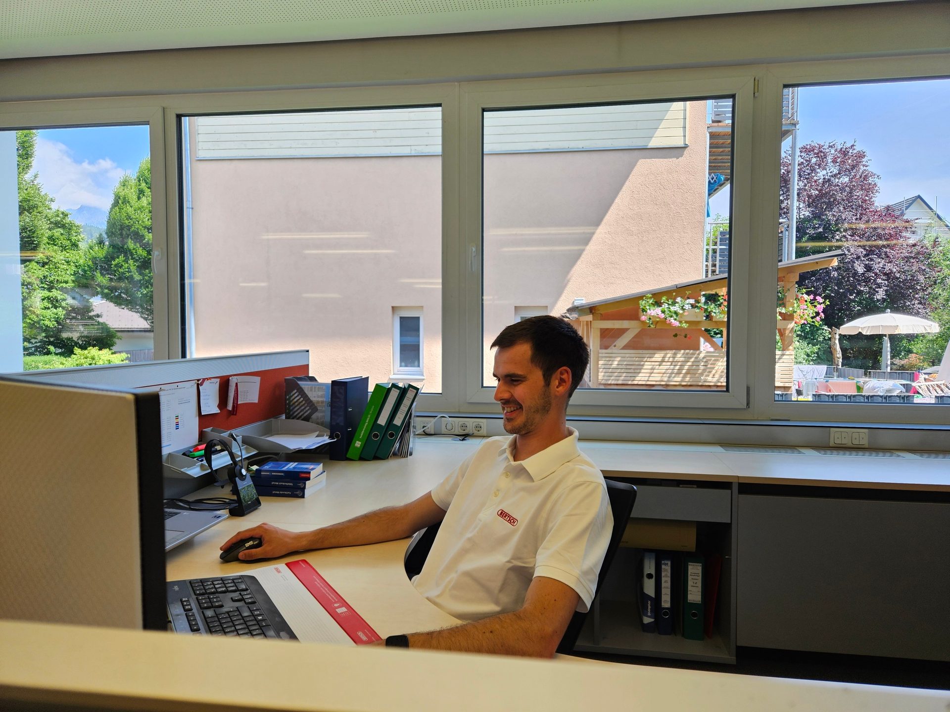 Smiling man at desk, looking at computer in office with large windows.