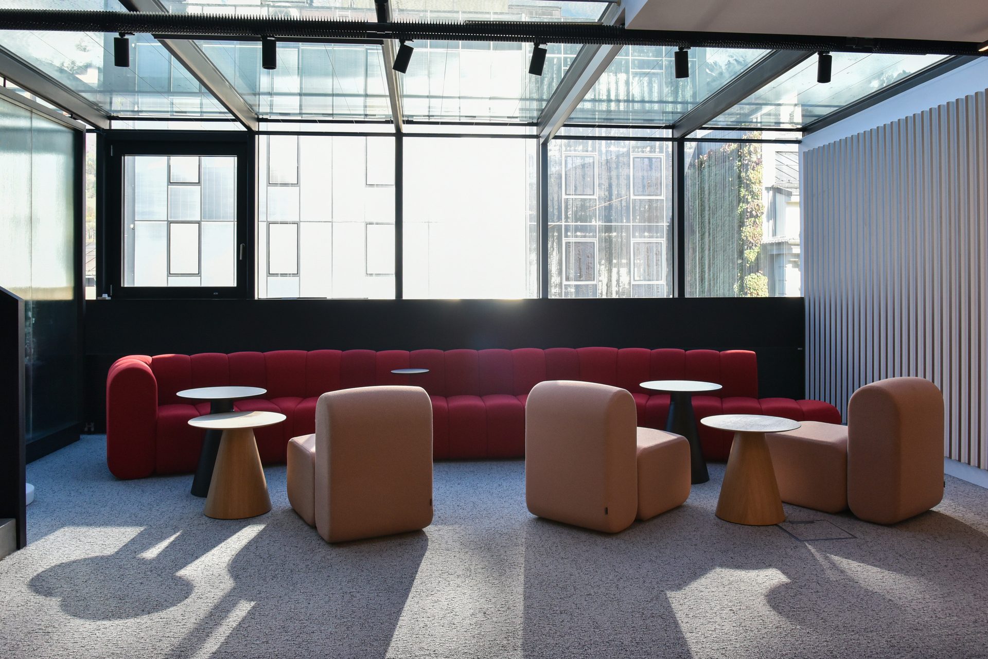 Modern lounge area with a long red modular sofa, light armchairs, and round tables under a glass ceiling.