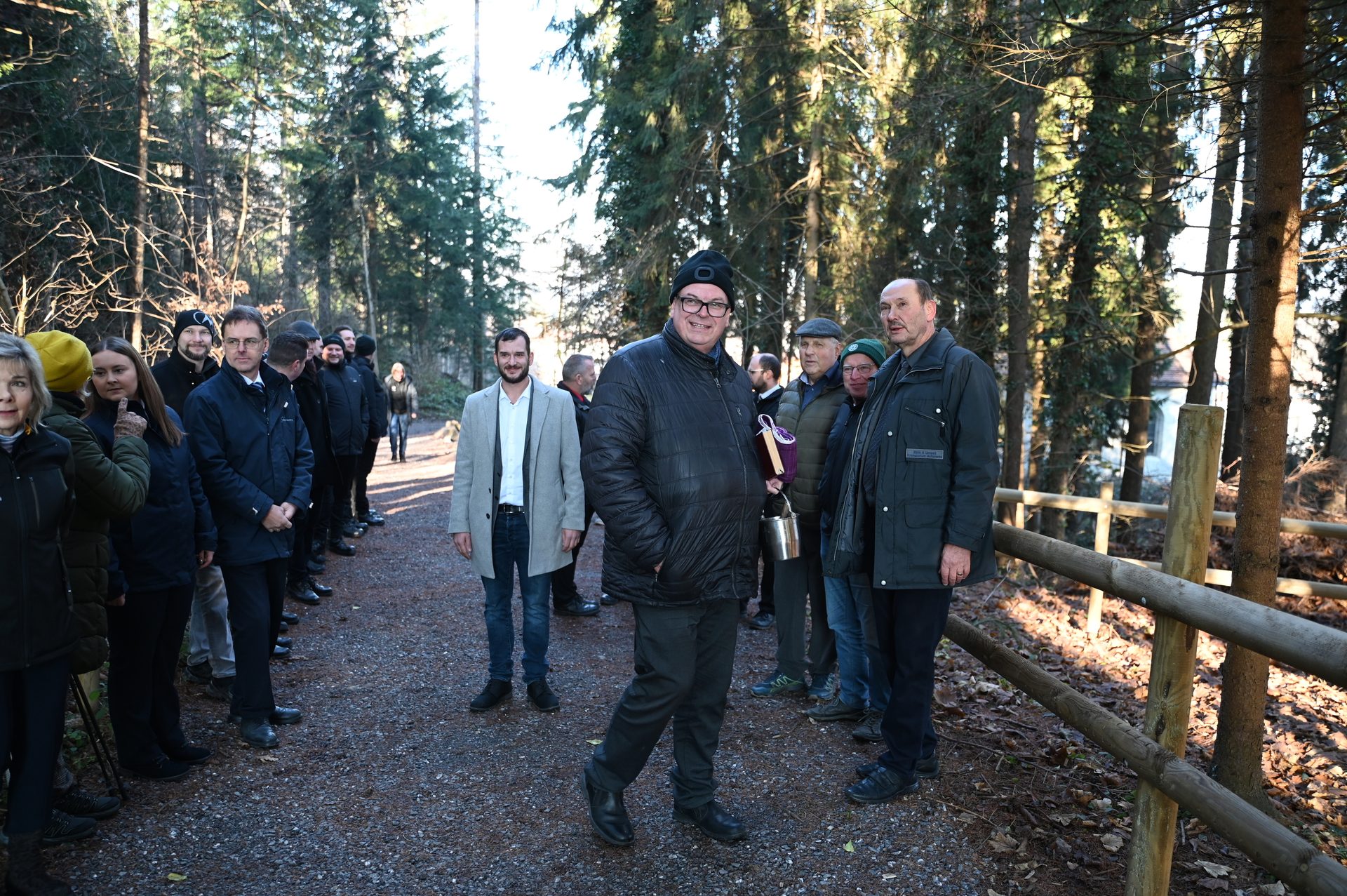 A smiling man in a black jacket stands among a group of people on a path in a sunlit forest.