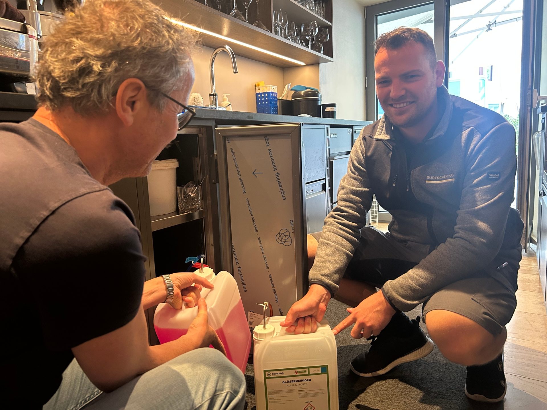 Two men discuss cleaning supplies in a kitchen, with one pointing to a glass cleaner.