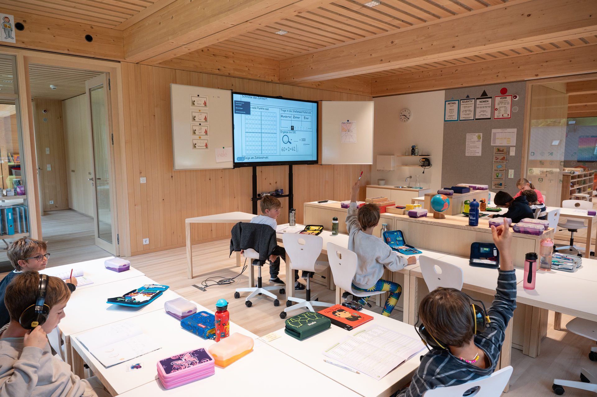 Children in a modern classroom with wooden decor, working at desks, some looking at a large screen.