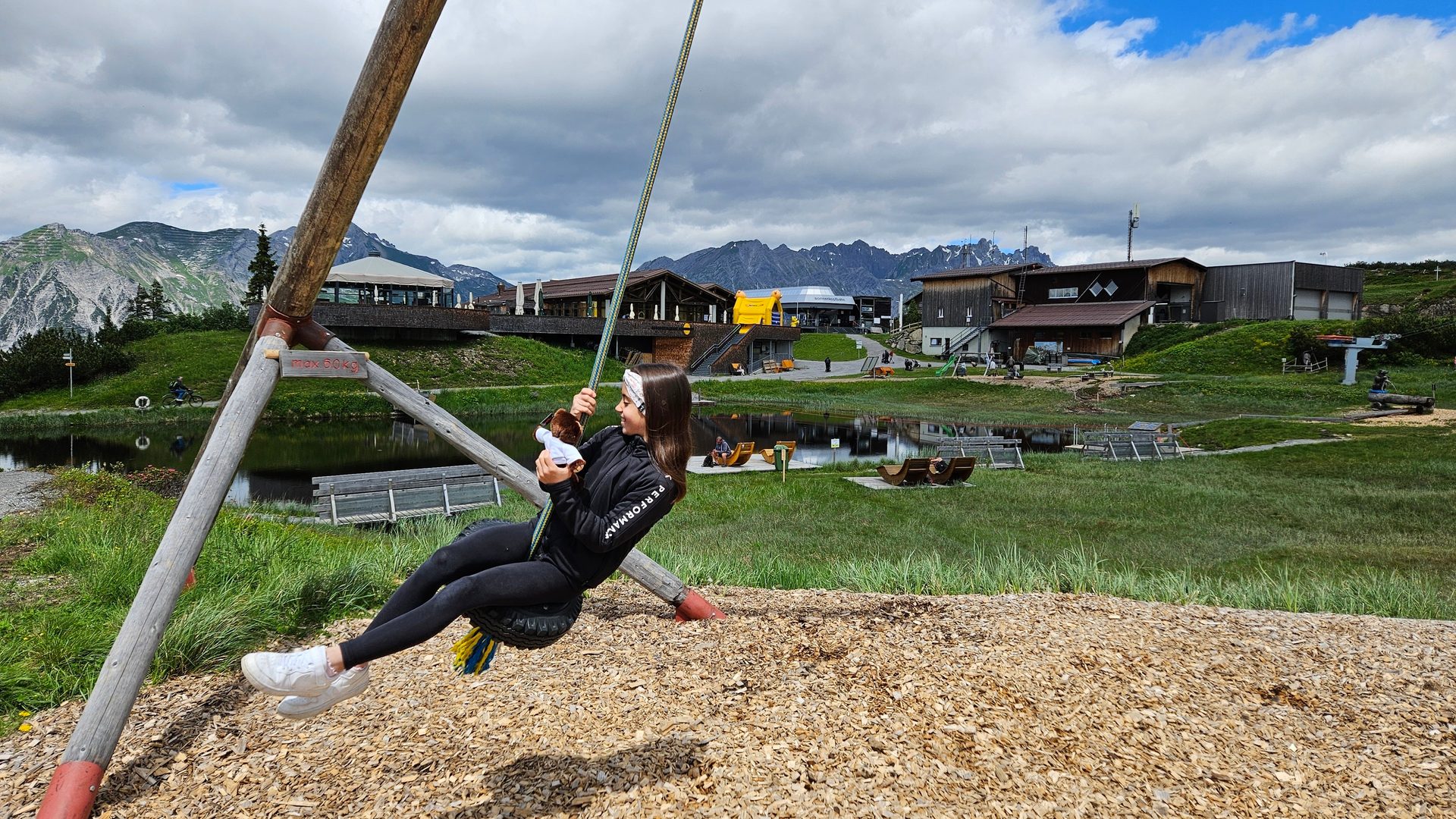 Girl swings on a tire swing at a mountain resort, surrounded by buildings, a lake, and green hills.