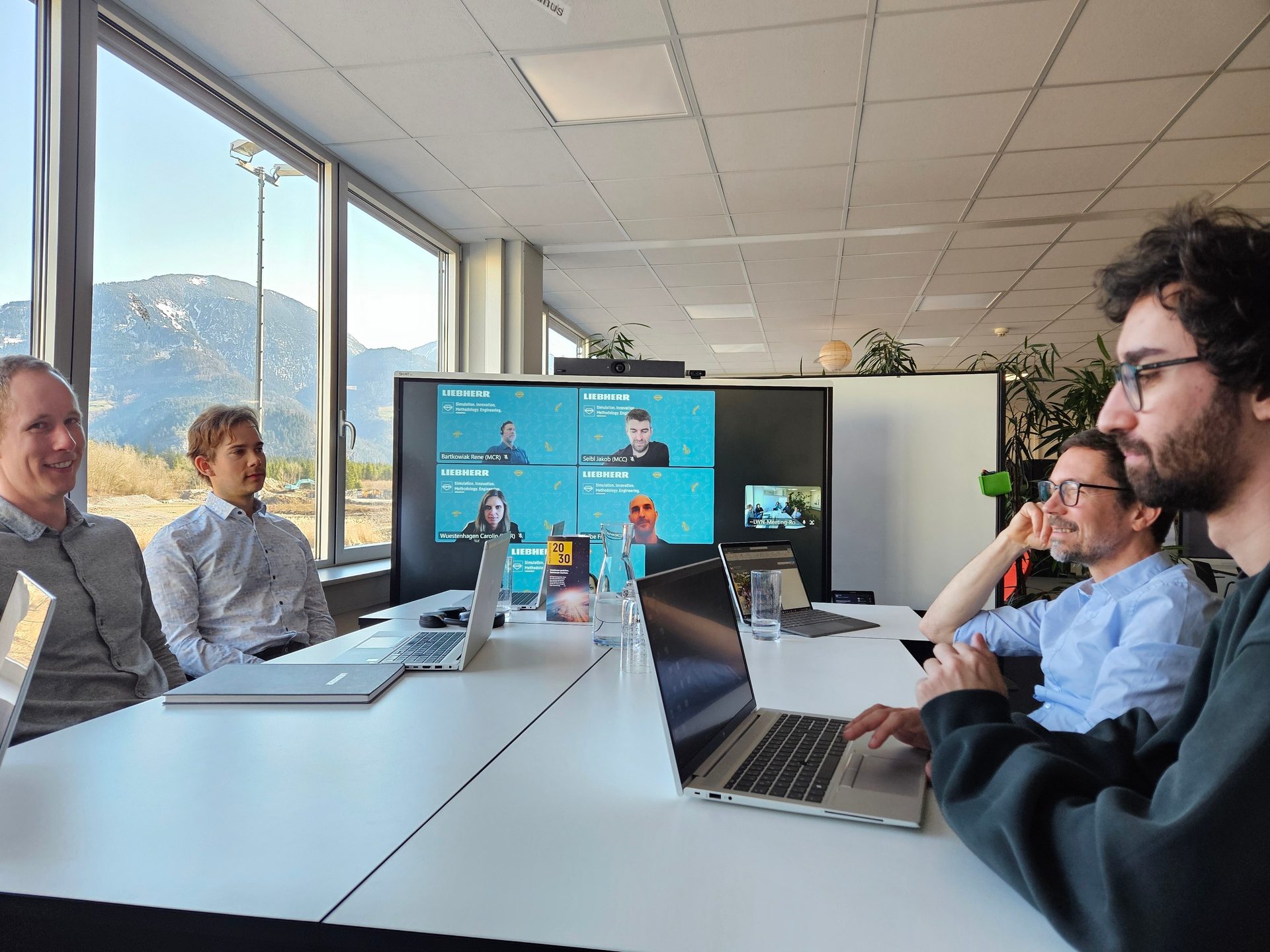 Men in a modern meeting room with a mountain view, engaged in a hybrid video conference.