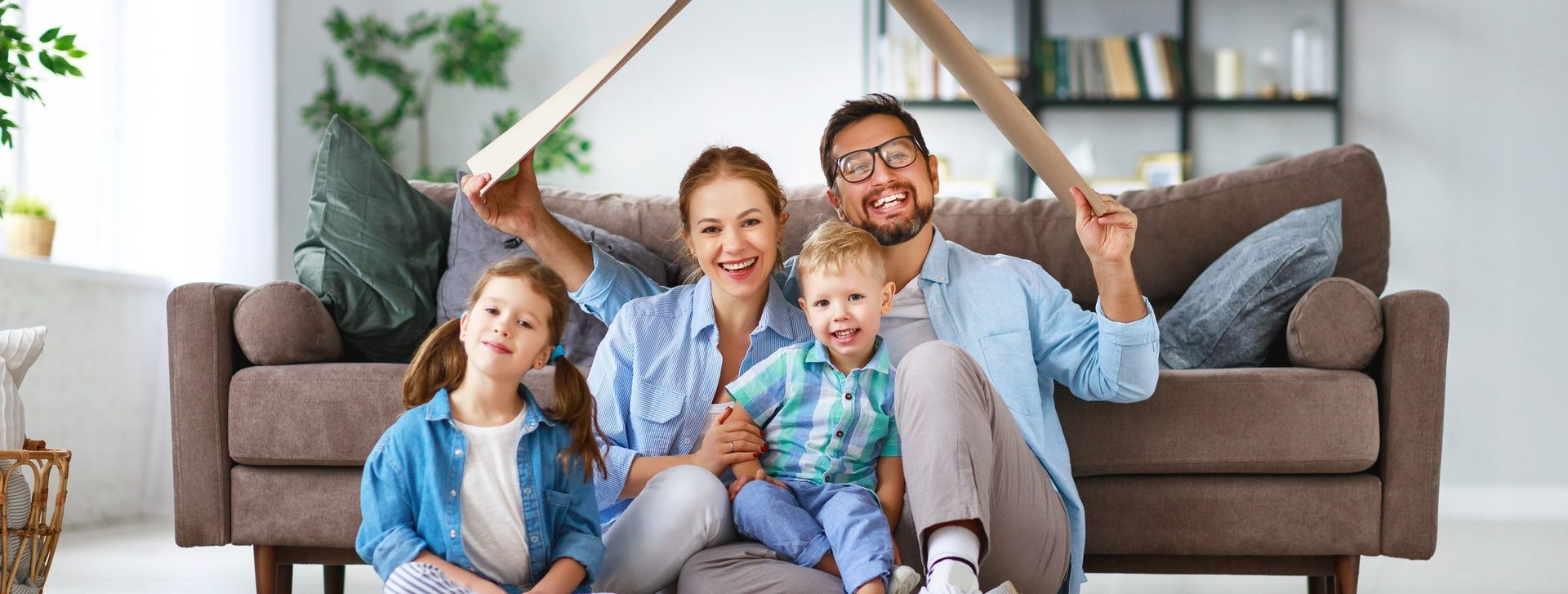 Smiling family of four, parents holding cardboard roof over kids, in front of sofa.