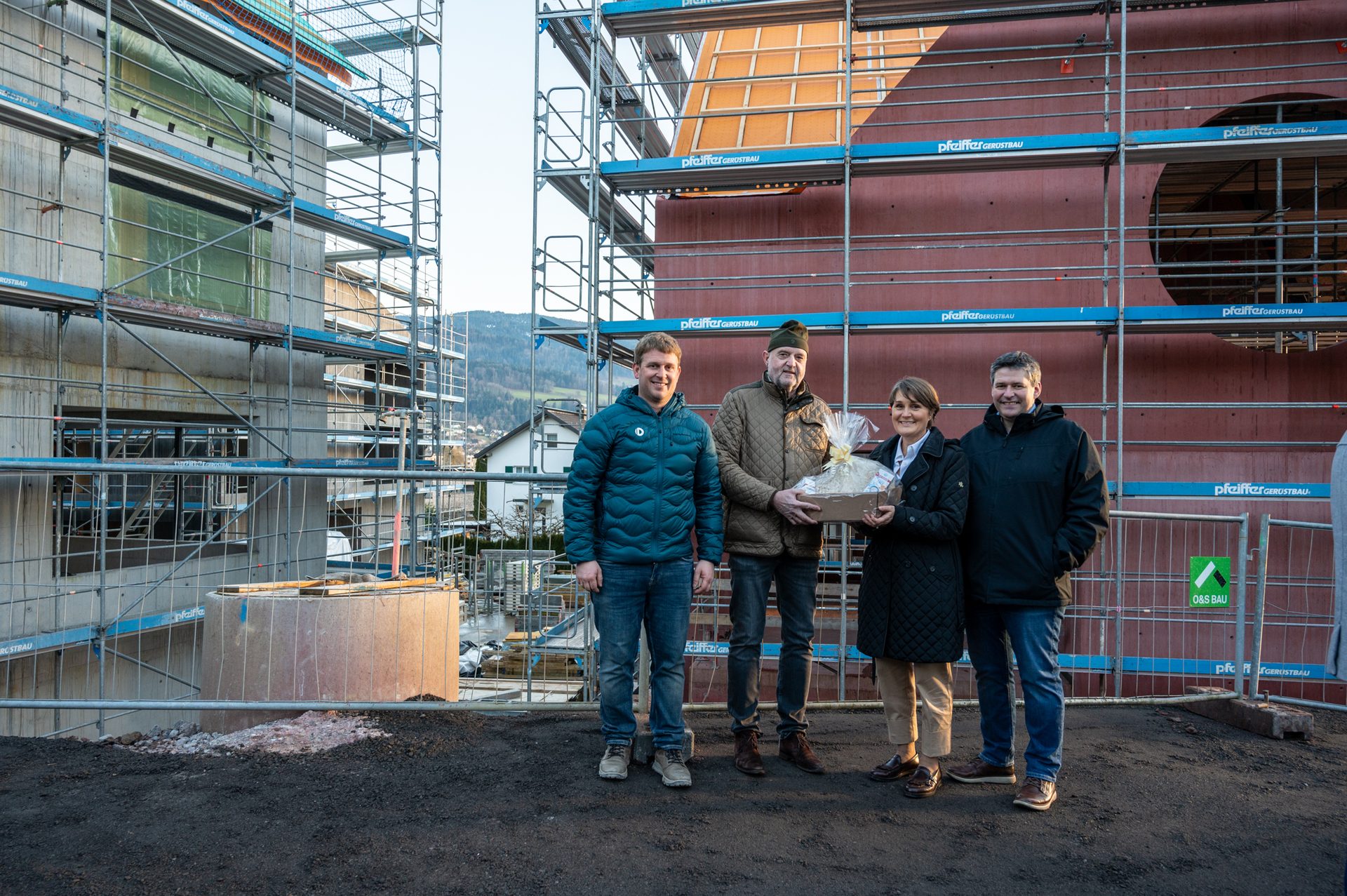 Four people on a construction site with scaffolding, one holding a gift basket.