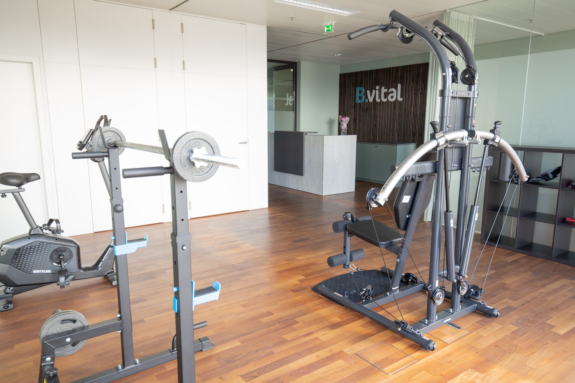Exercise bike, barbell rack, and multi-gym in an office reception area with 'B.vital' logo.