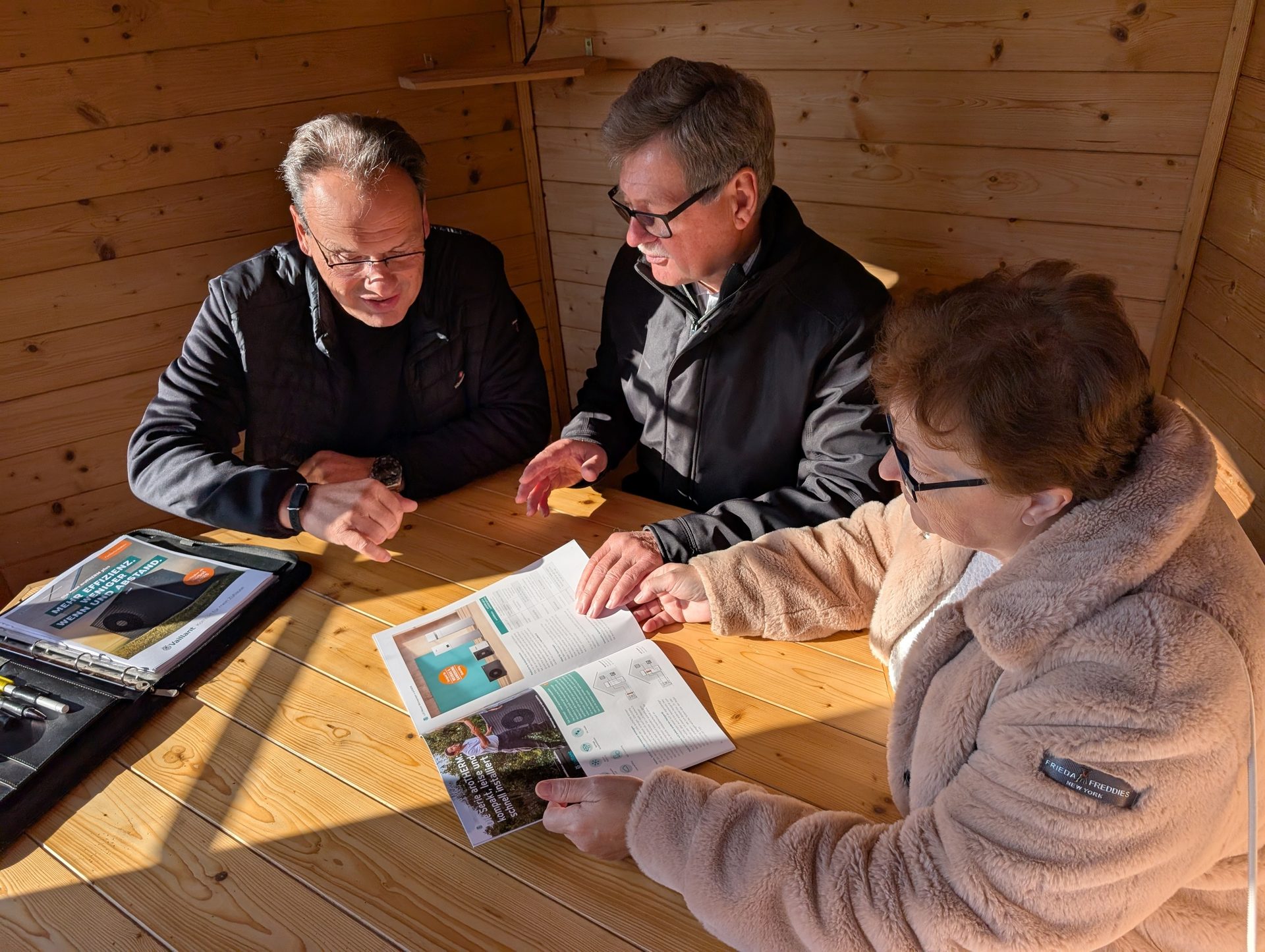 Three people discuss documents on a wooden table, one holding an open brochure.