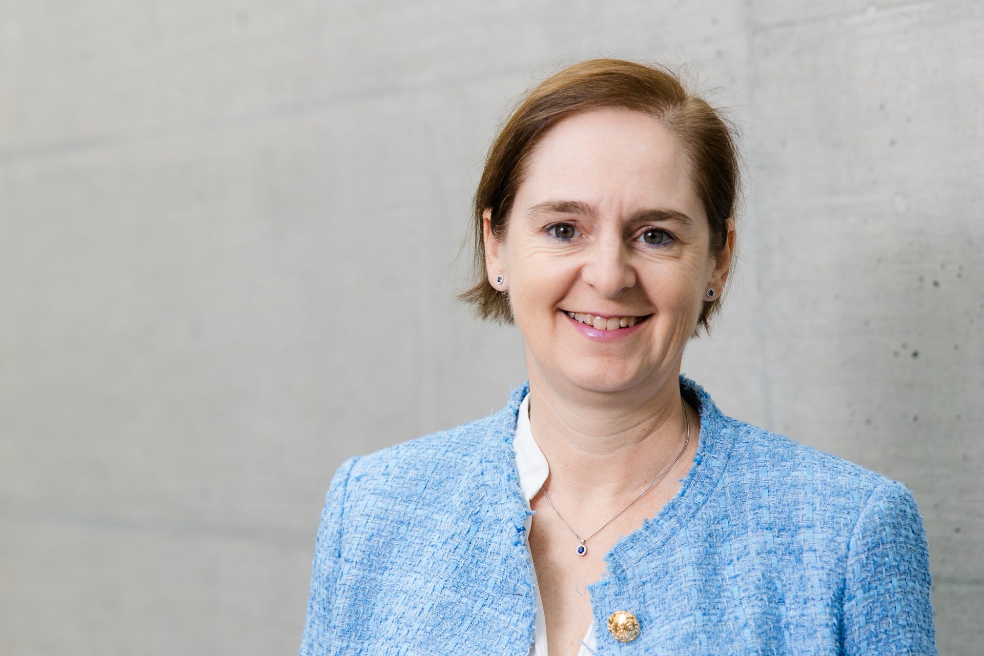 Smiling woman in a blue textured jacket, with short brown hair and blue jewelry.