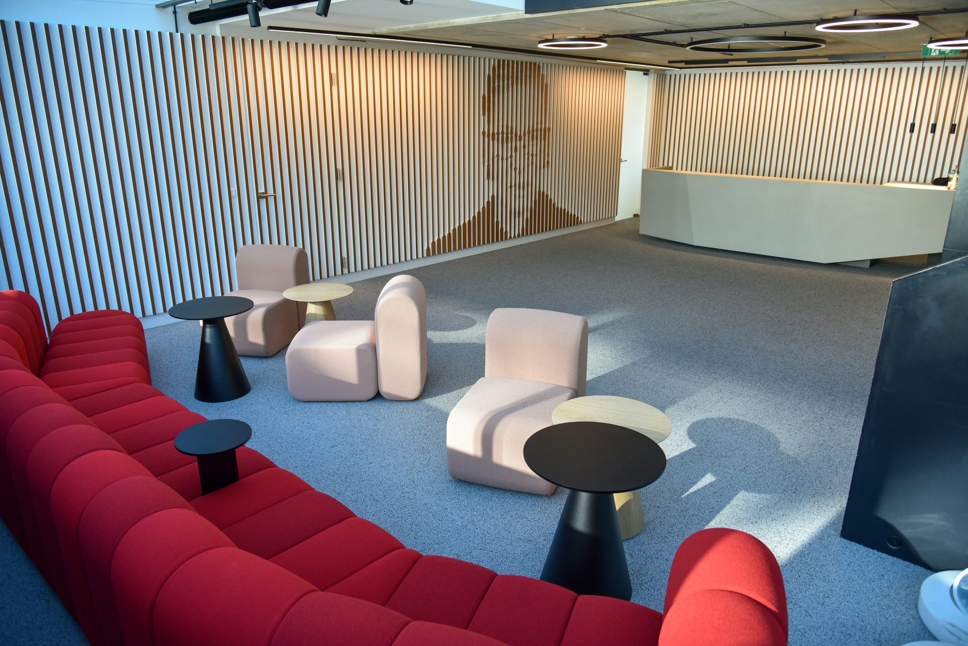 Modern reception lounge with red sofa, light pink chairs, small tables, and slatted wall with face art.