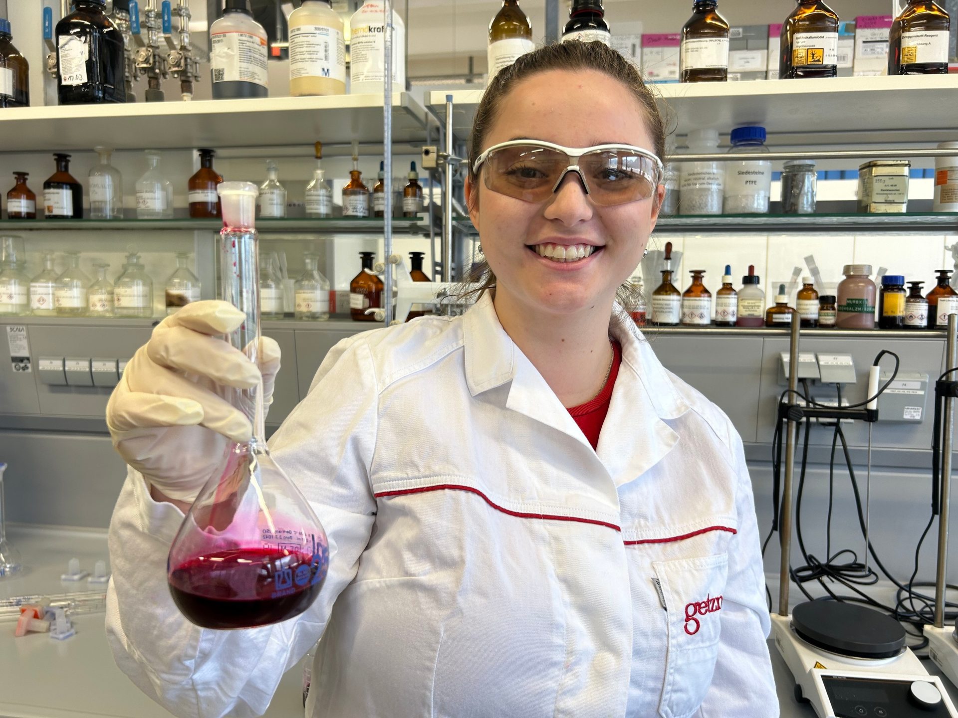 Smiling scientist in lab coat and glasses holding a red liquid flask in a lab.