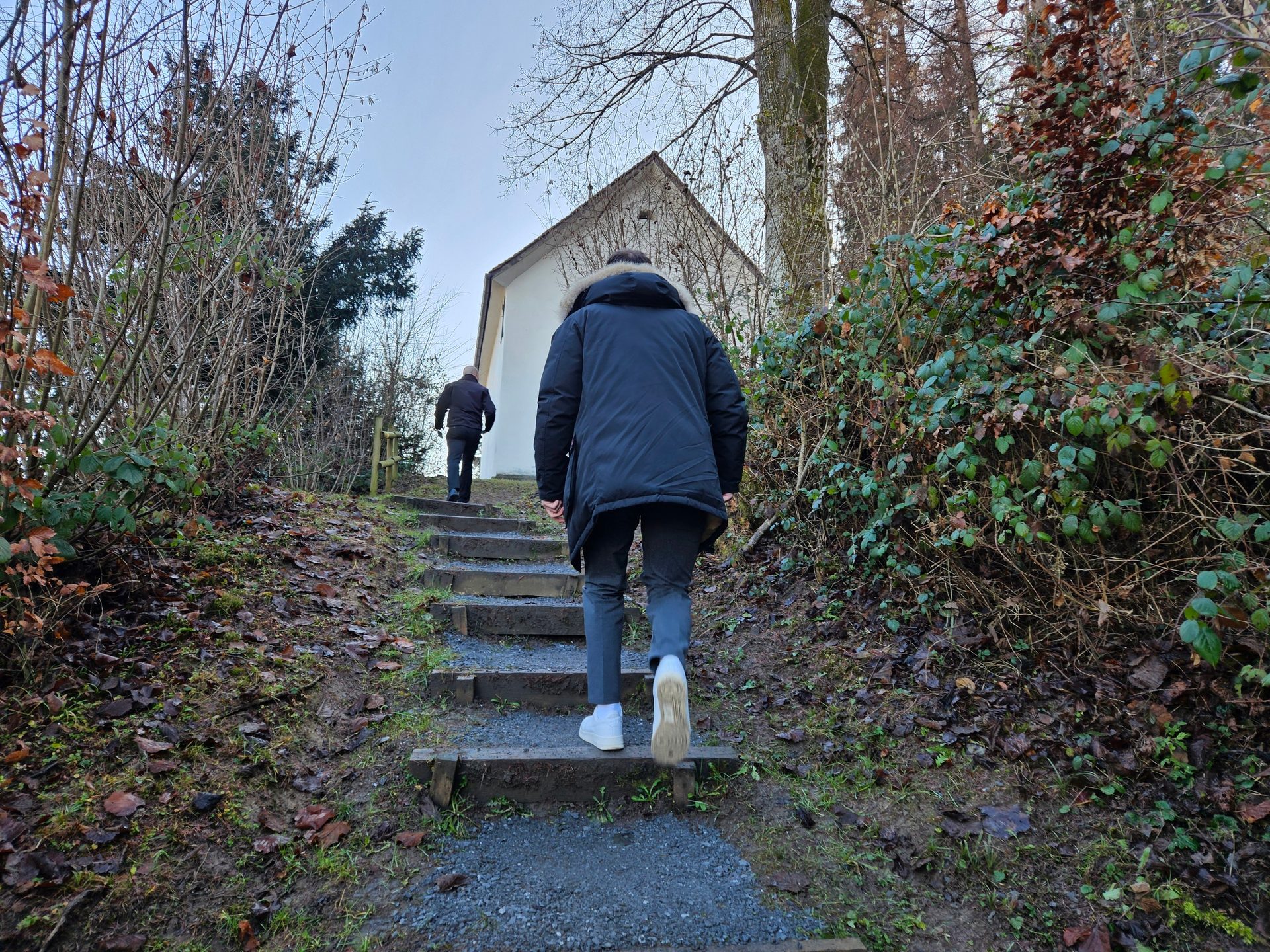 Two people ascend wooden steps toward a white building, surrounded by bare trees and muddy ground.