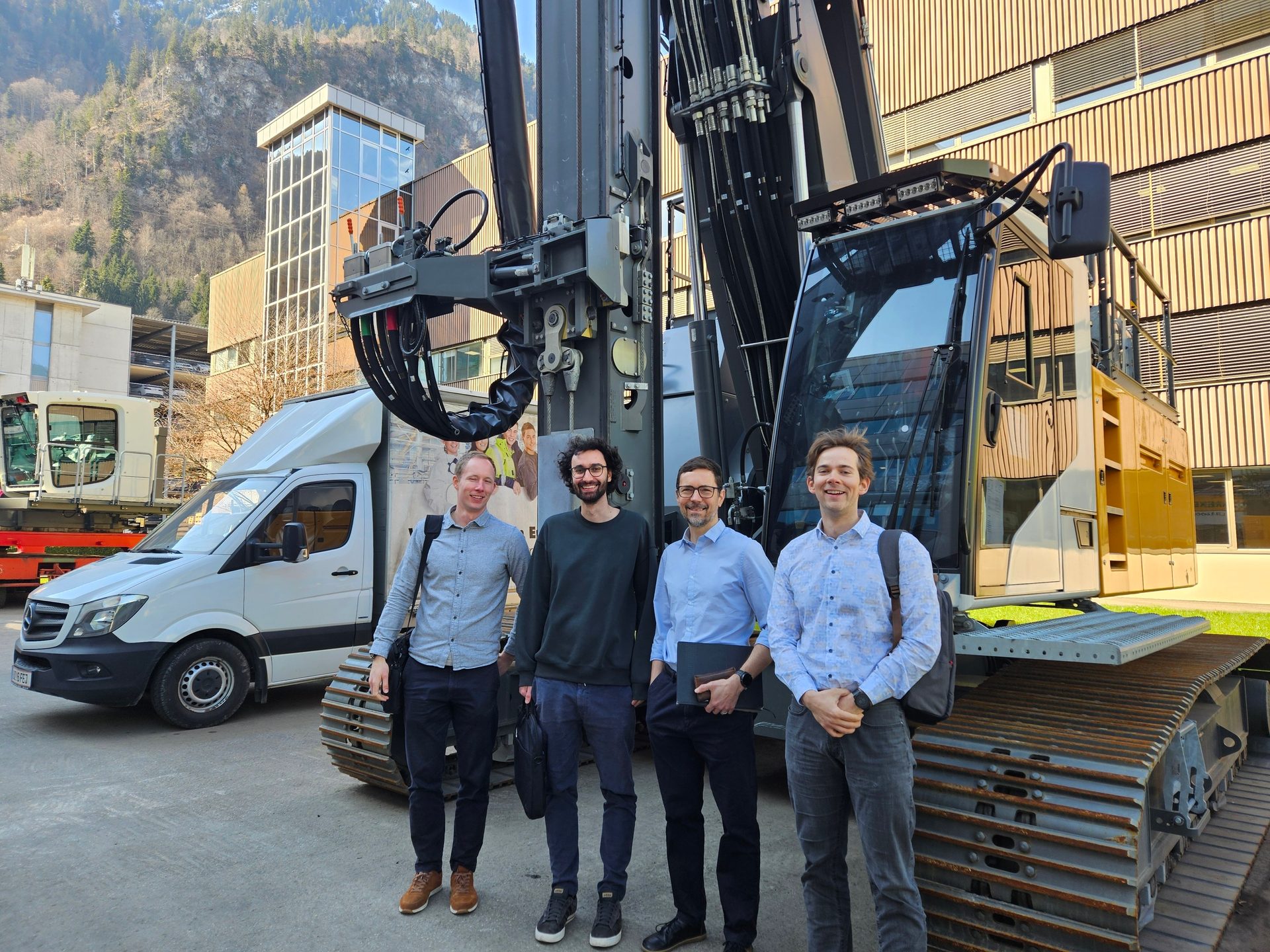 Four men stand by a large construction rig and a white van, mountains in the background.