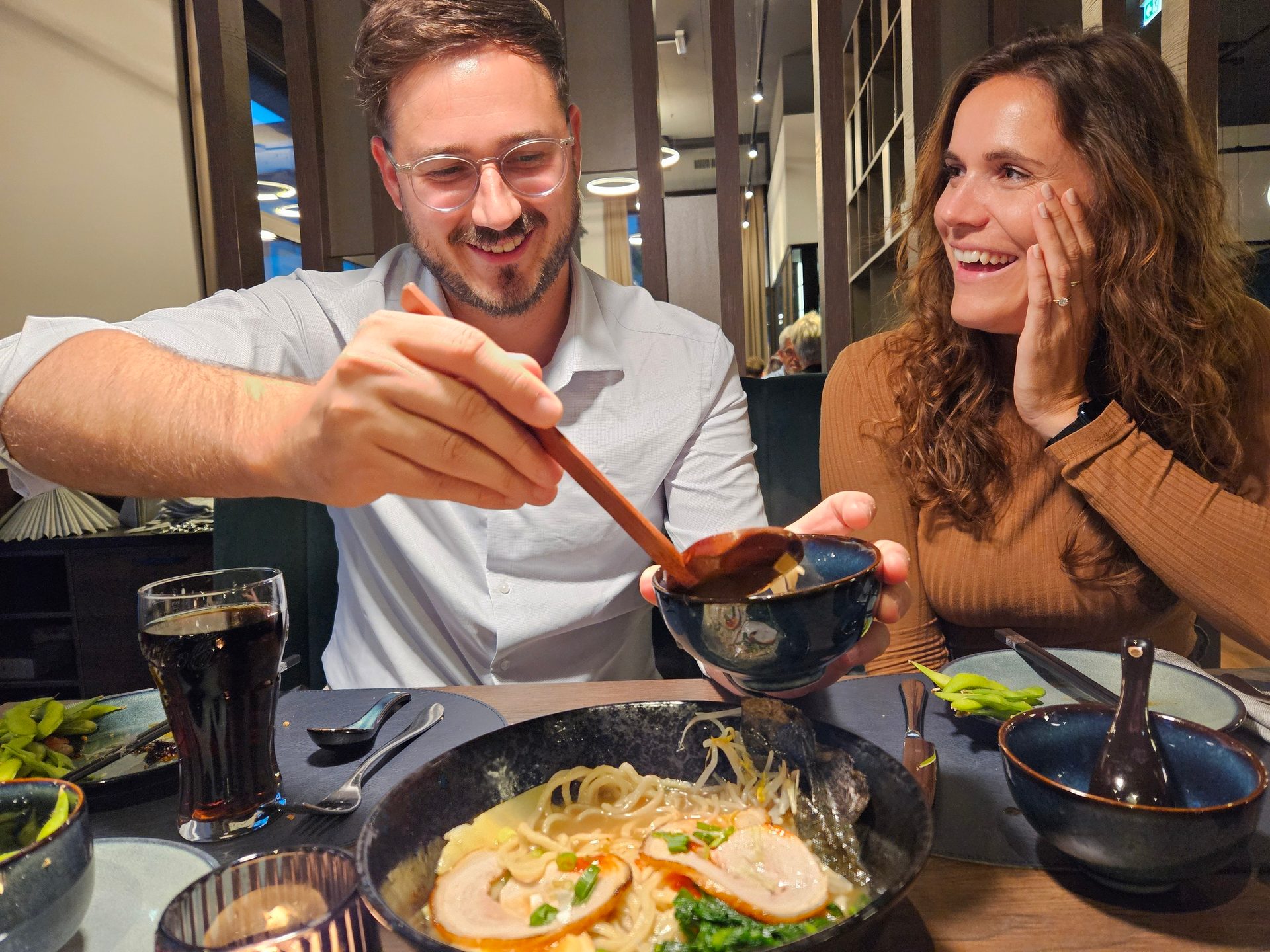 Smiling man serving ramen to a happy woman at a restaurant table.