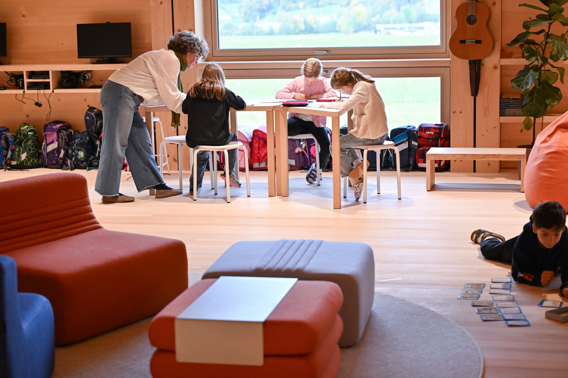 An adult helps children writing at a table; another child plays on the floor in a bright room.