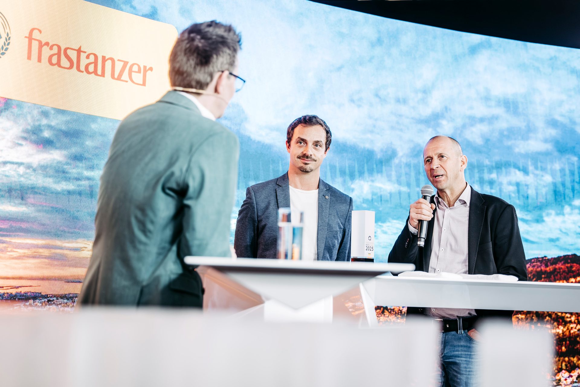 Three men on a stage, one speaking into a microphone, with a large screen displaying a landscape.