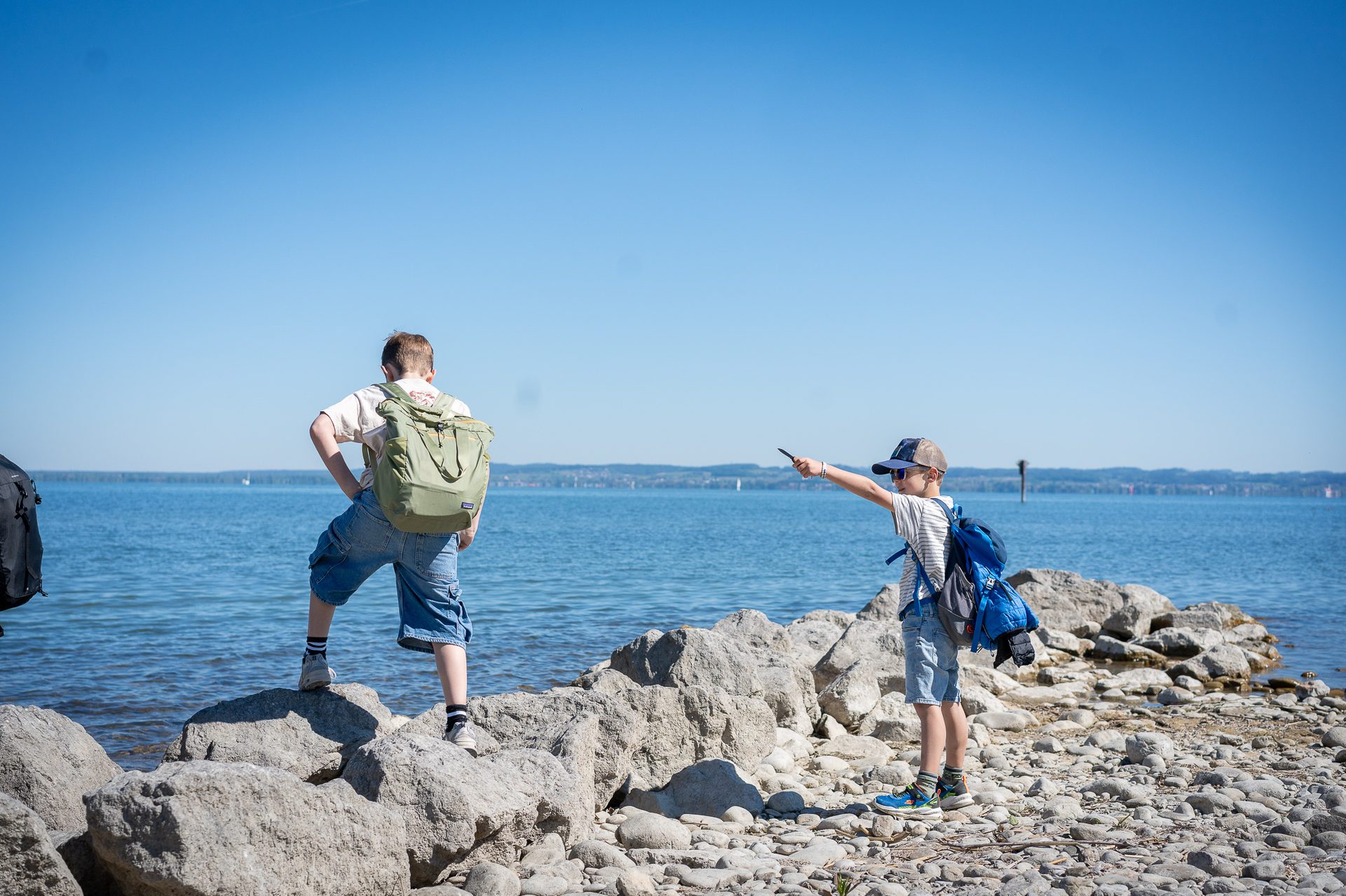 Two boys with backpacks on a rocky lake shore, one pointing across the water.
