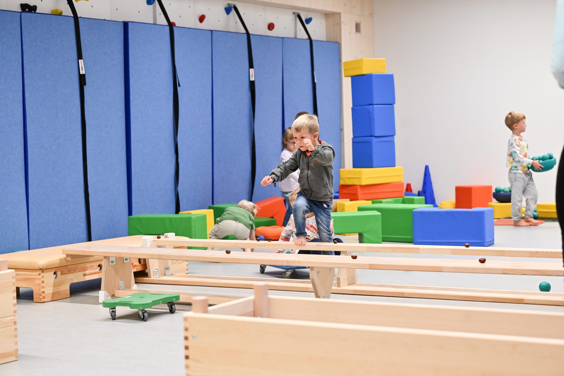 Kids play in a bright indoor gym: a boy balances on a wooden beam, others interact with colorful blocks.