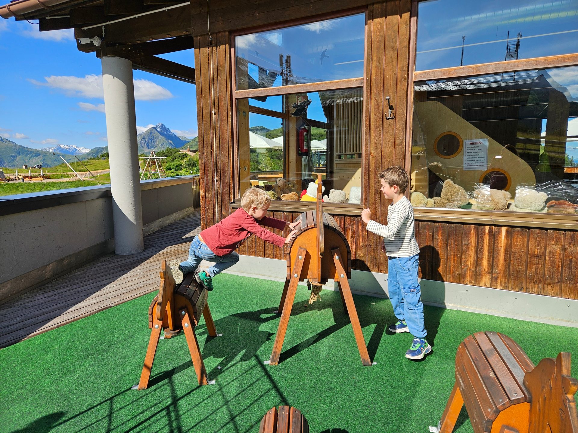 Two boys play on wooden barrel toys on a green outdoor surface with mountains in the background.