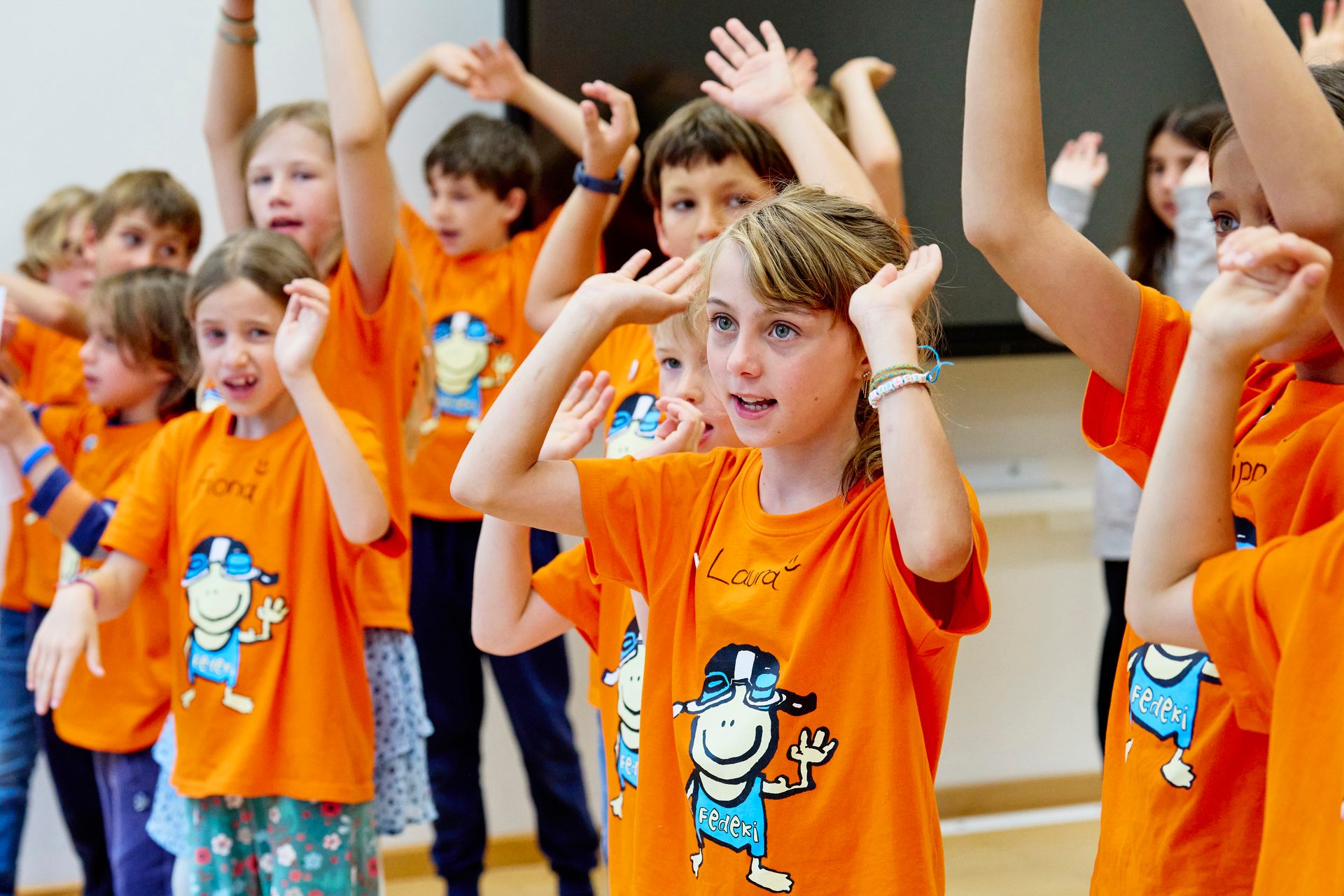 Group of children in orange shirts enthusiastically participating with raised hands in an indoor setting.