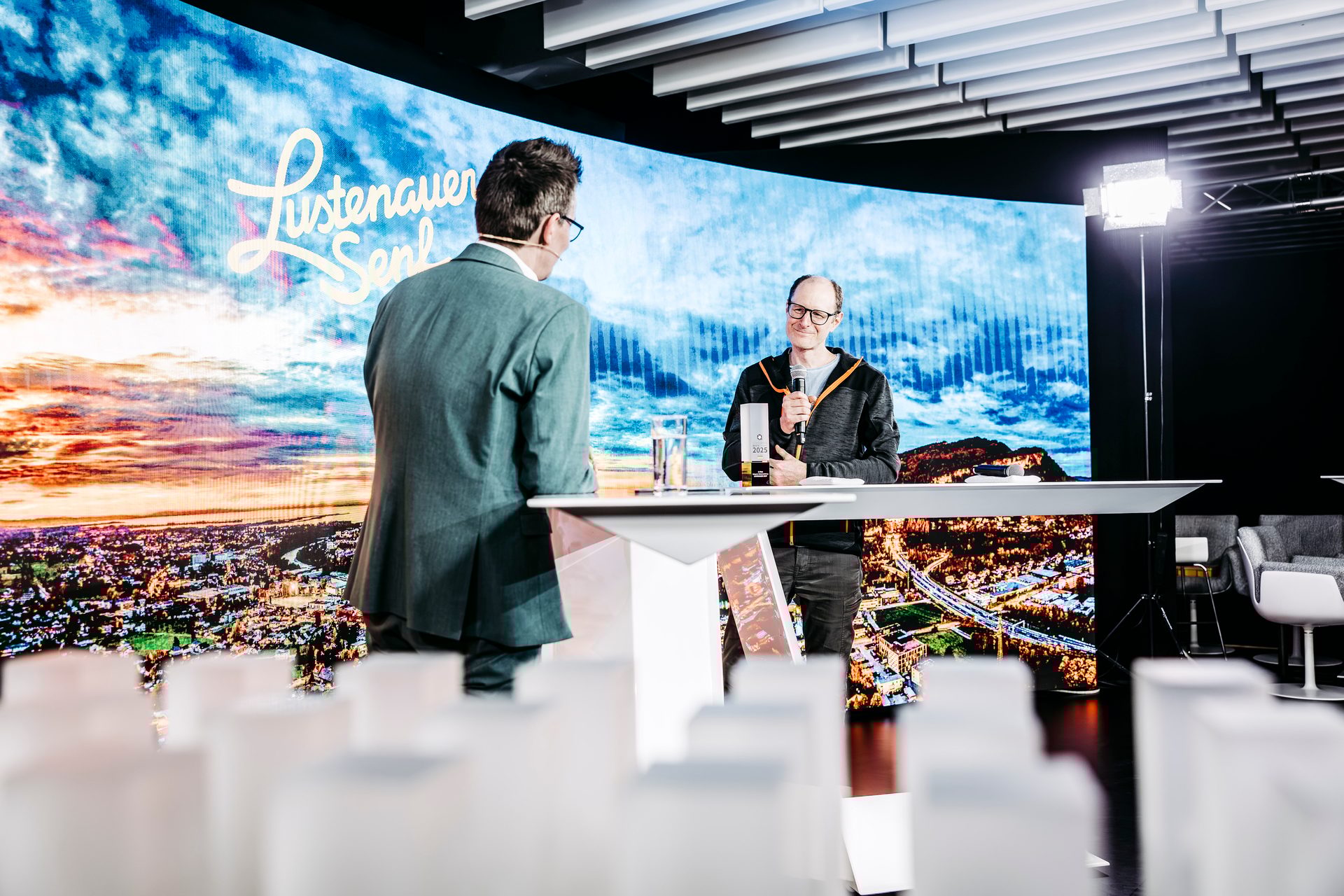 Two men, one speaking into a mic, on a stage with a large LED screen displaying a city and sky.