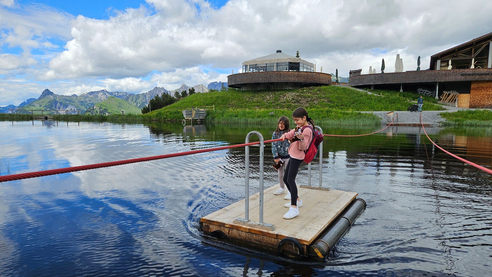 Two girls on a raft crossing a mountain lake, with a building on a hill in the background.