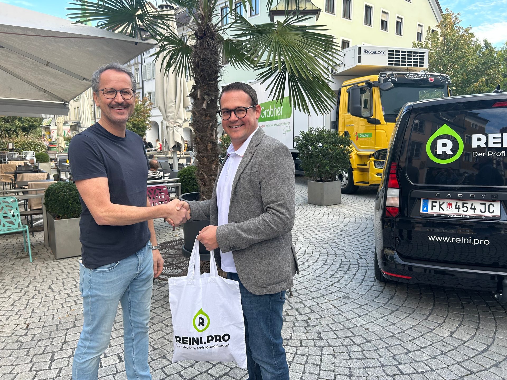 Two men shaking hands, one holding a "Reini.pro" bag, with delivery vehicles in the background.