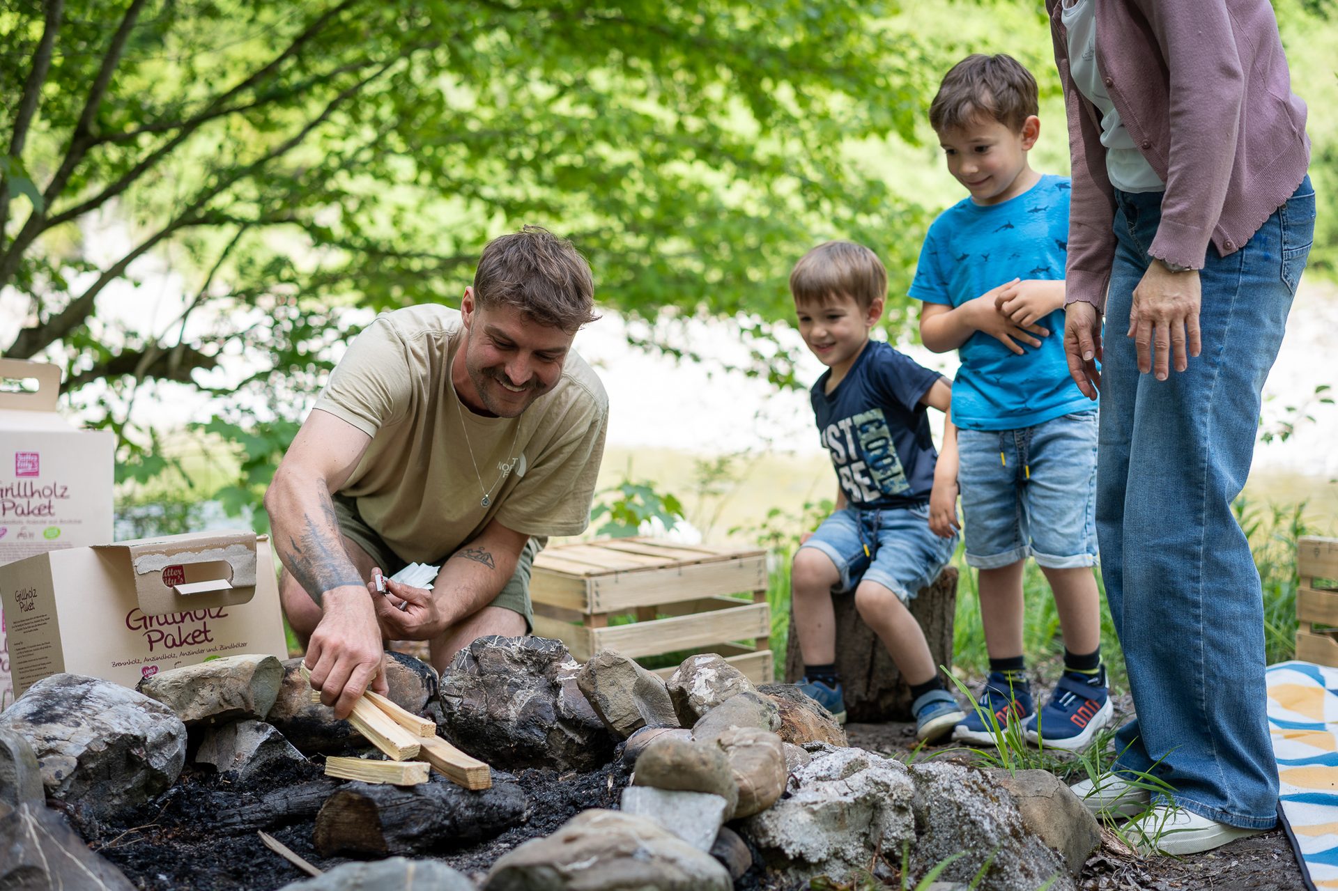 A man and two young boys preparing a campfire outdoors, with the man placing small wood pieces.