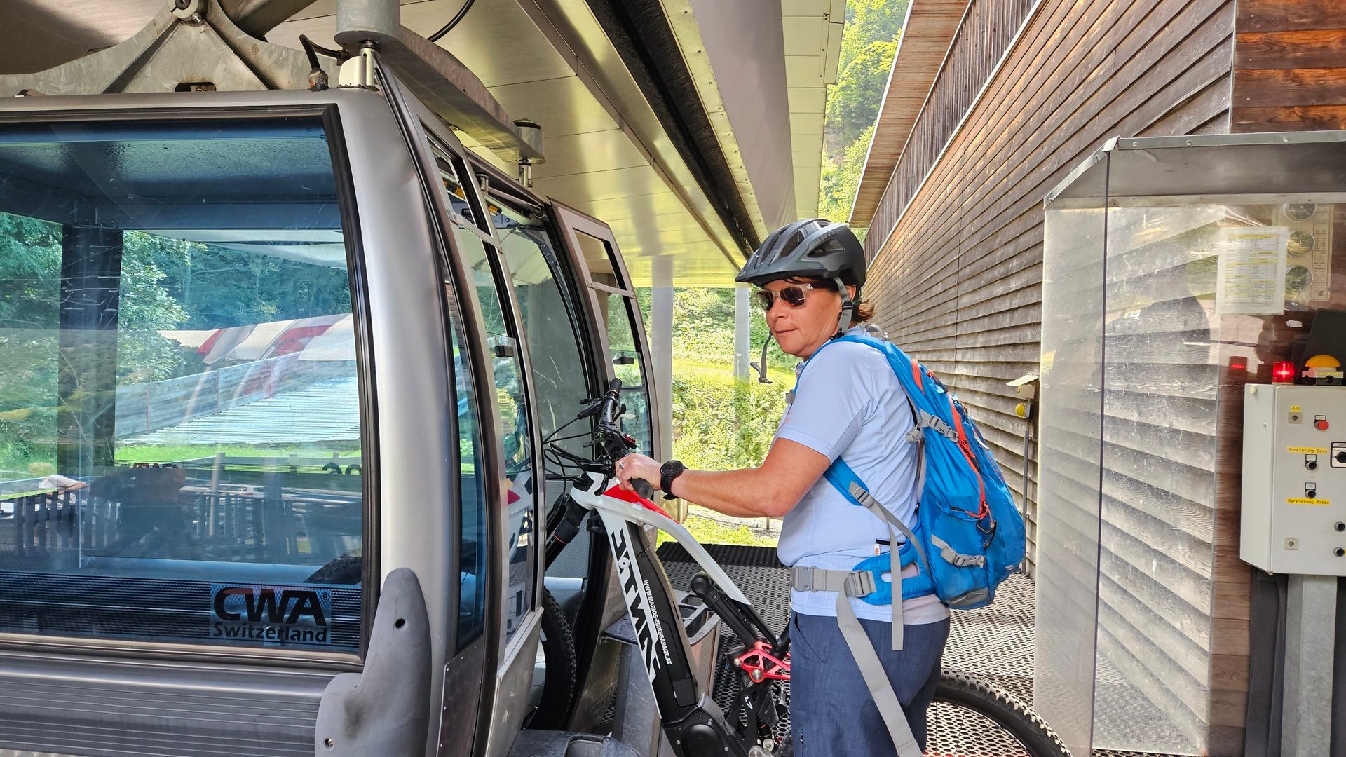 A person in a helmet and backpack is loading a mountain bike onto a gondola lift at a station.