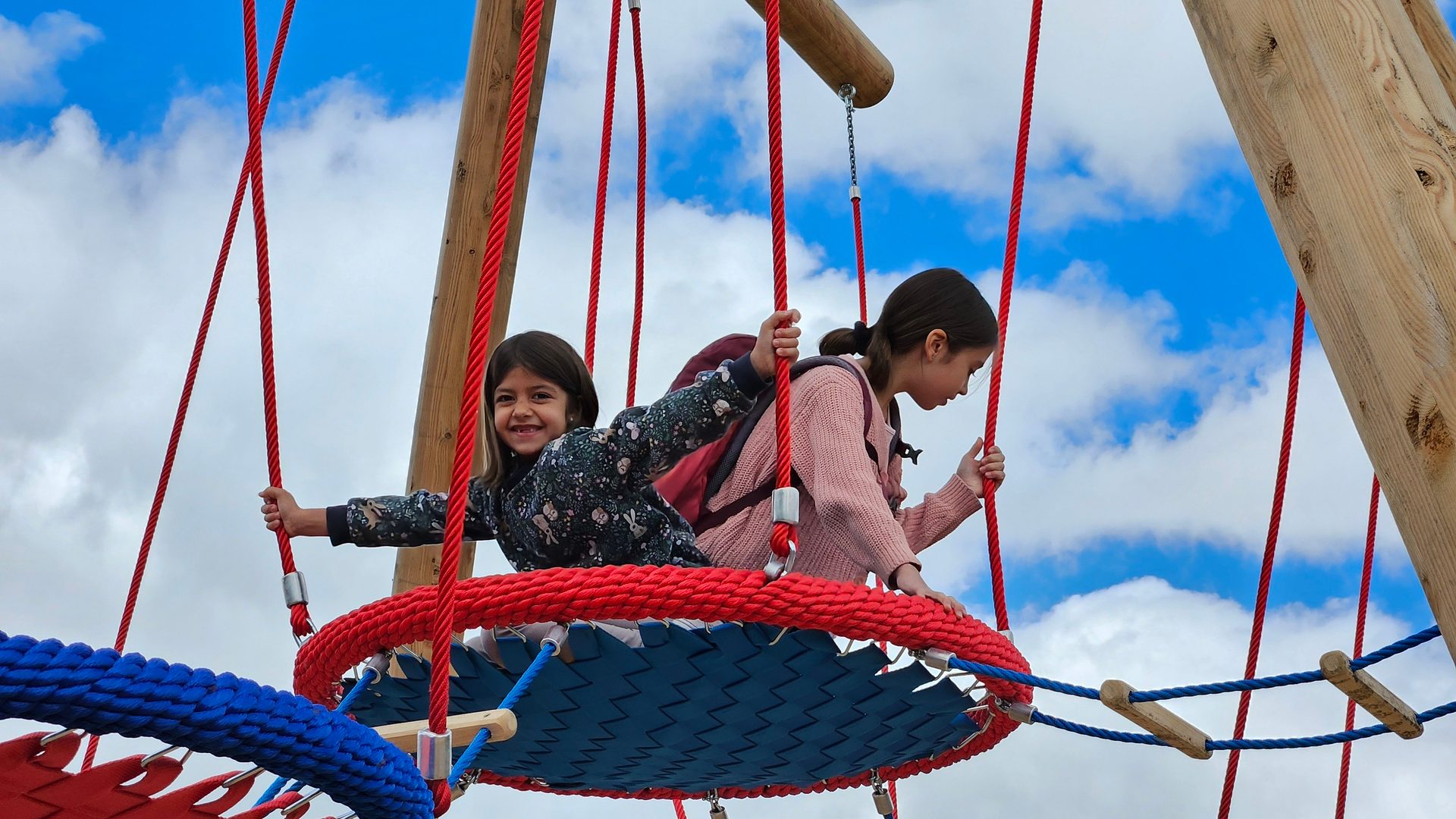 Two girls smile and play on a colorful rope swing under a blue sky with white clouds.