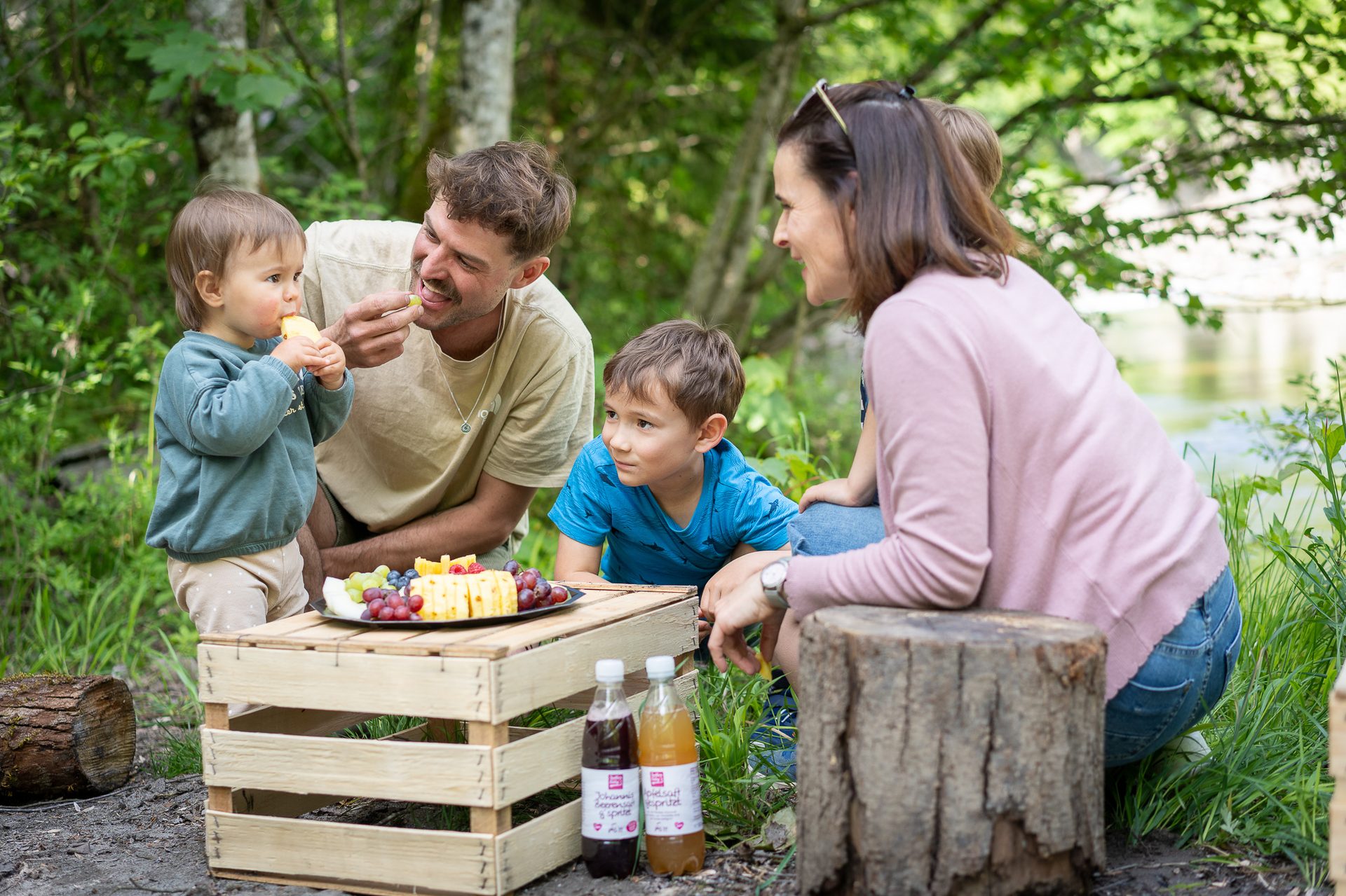 A family (parents and two children) enjoys a picnic with fruit and juice outdoors in a lush green forest.