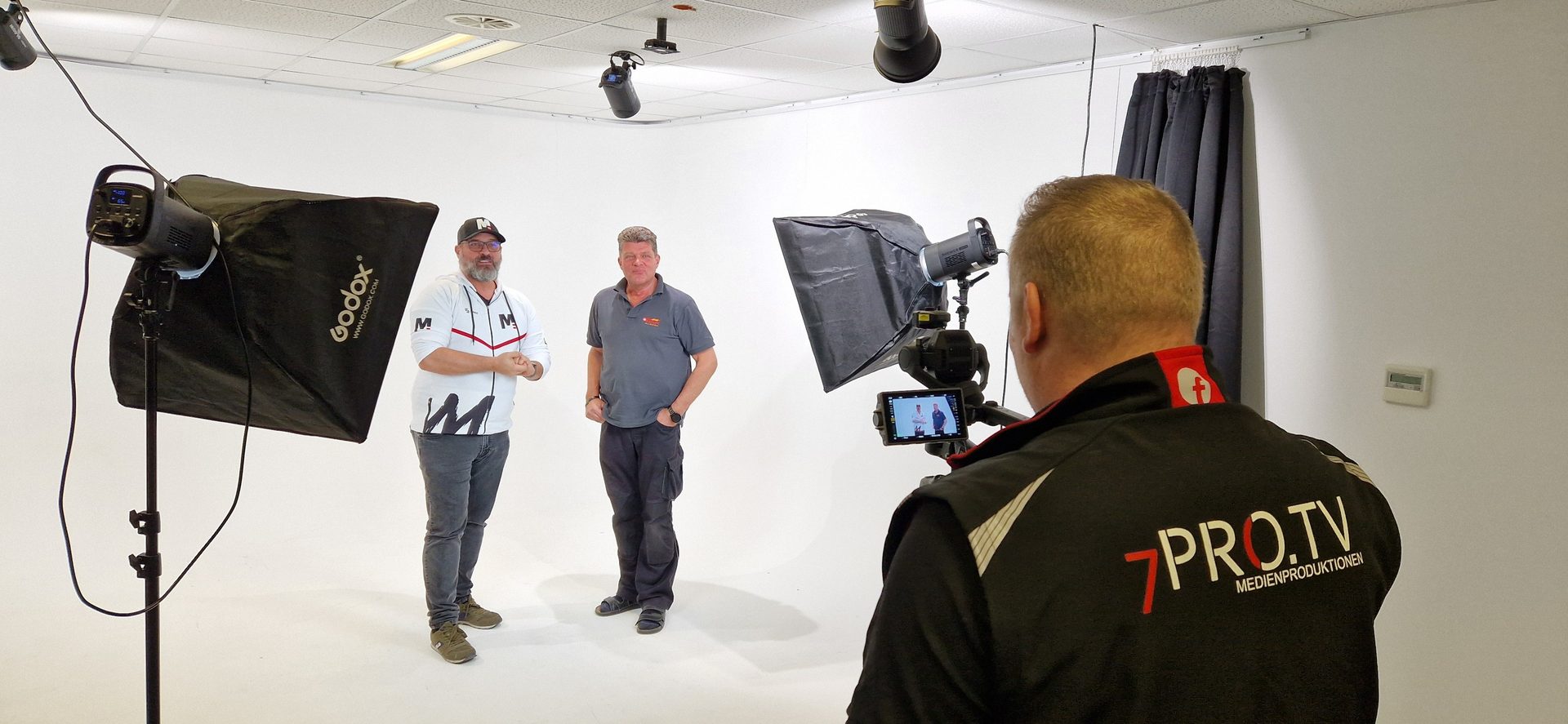 Man filming two men in a white studio with video camera and softboxes.