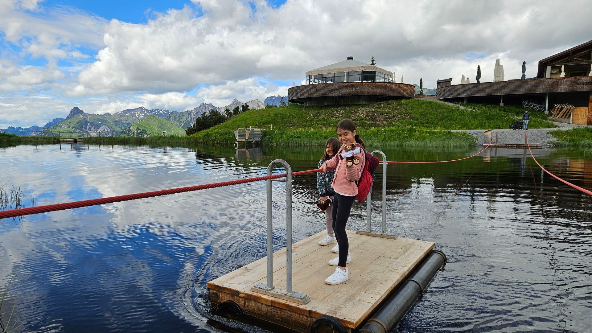Two girls on a wooden raft in a lake, with mountains and buildings in the background.