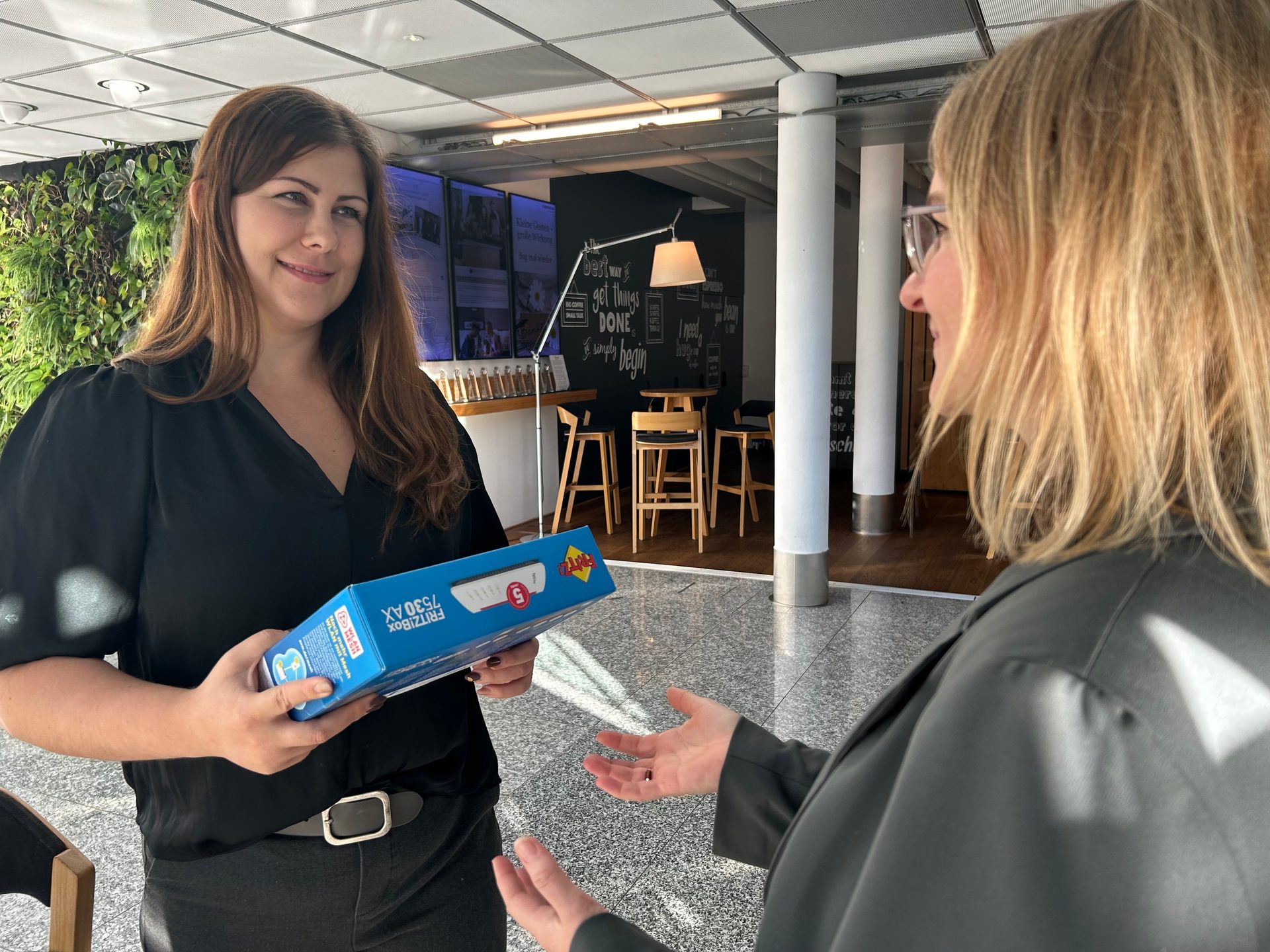 A smiling woman hands a blue FRITZ!Box to another woman in an indoor setting.