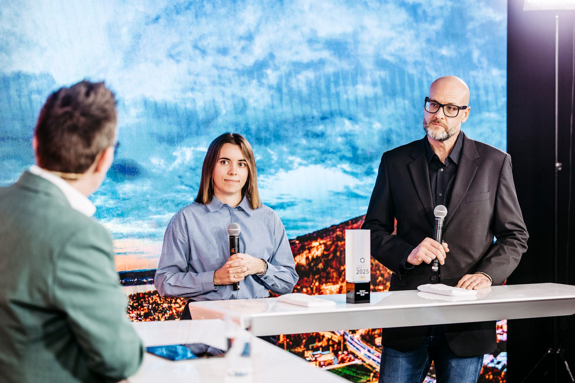Three speakers at a panel, two holding mics, against a large screen background.