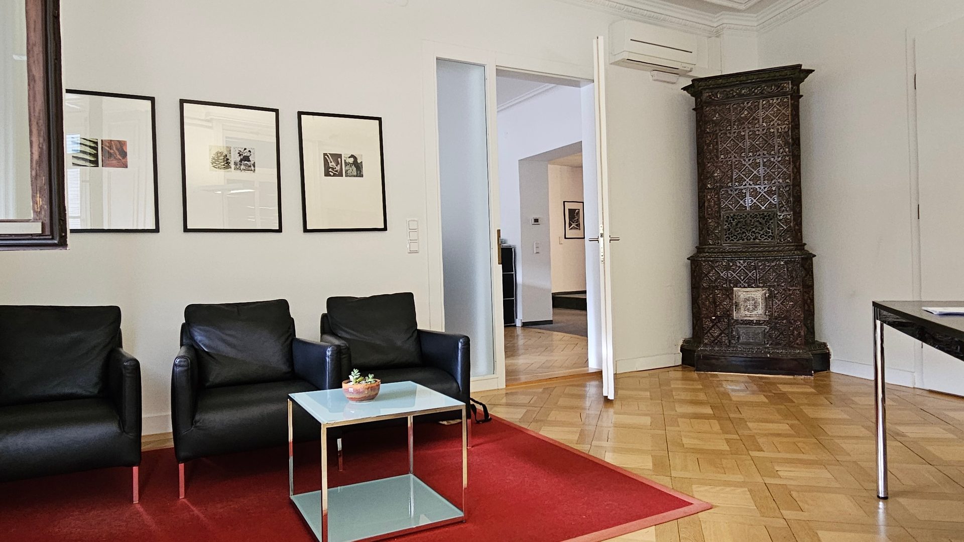 A waiting room with black armchairs, a glass table, framed pictures, a red rug, and an ornate tiled stove.