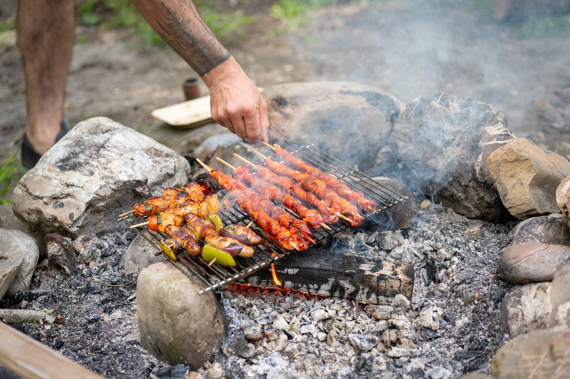 Man grilling chicken and vegetable skewers over a campfire, with smoke rising from the coals.