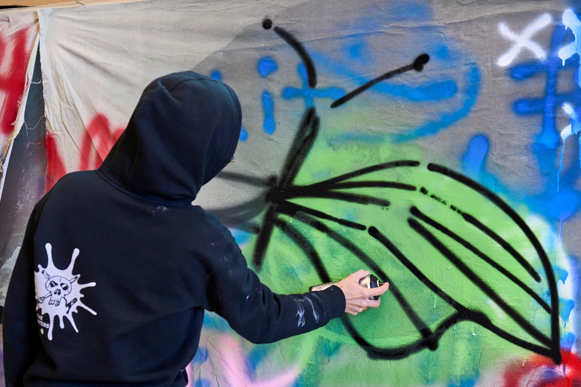 A person in a black hoodie spray painting a large green and black graffiti design on a canvas.