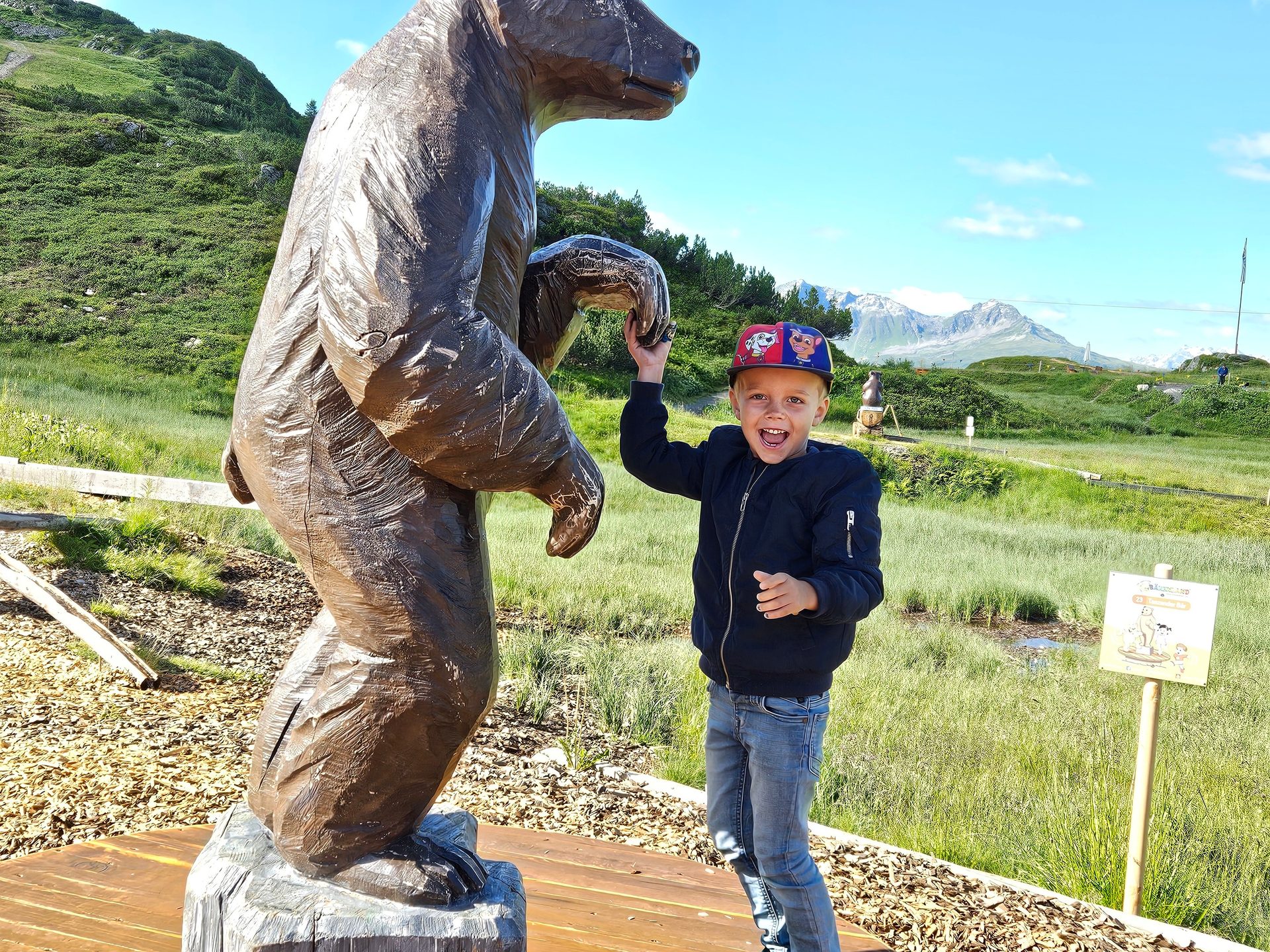 A happy boy poses next to a wooden bear statue in a sunny mountain landscape.
